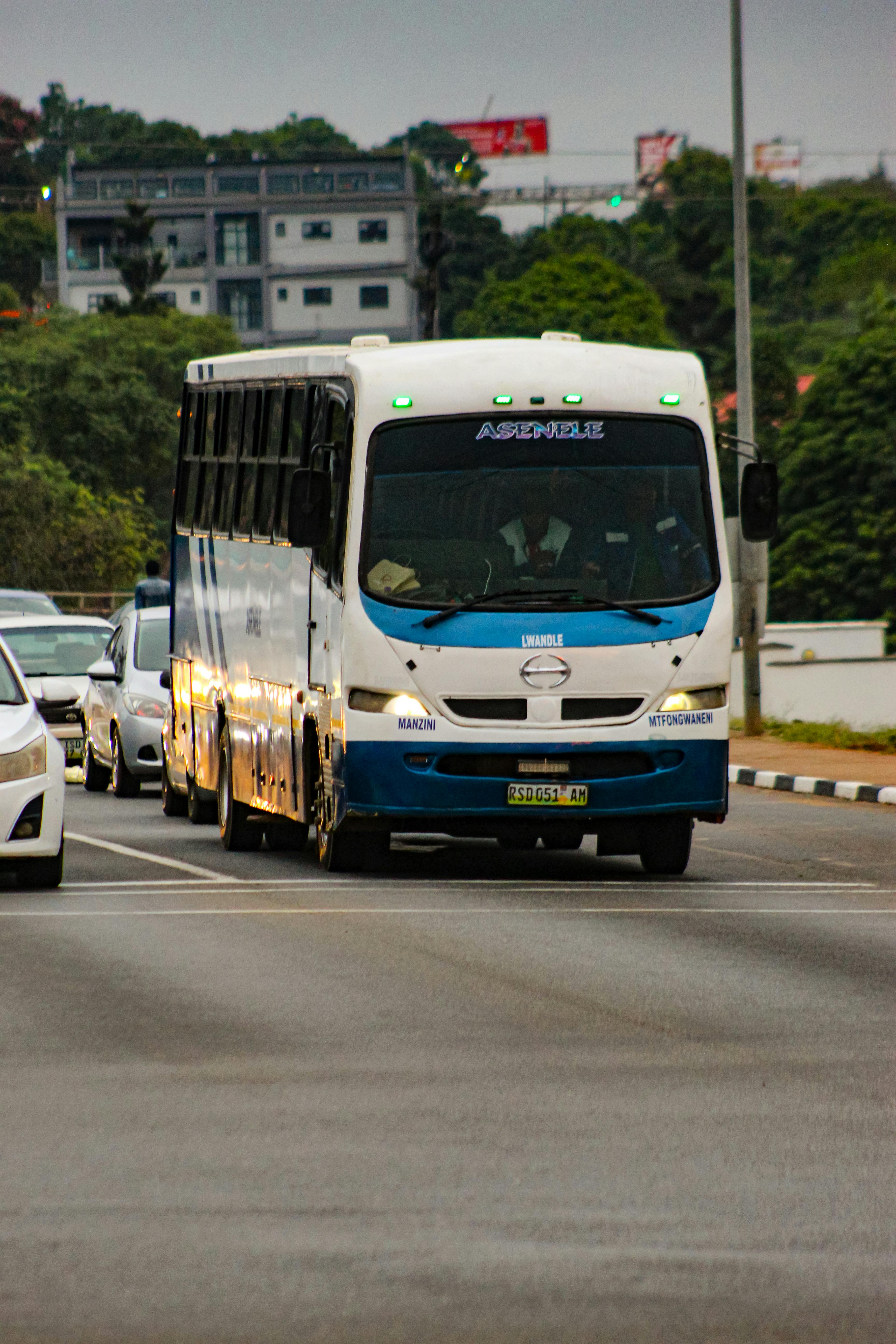 A white minibus navigates through city traffic, surrounded by greenery and modern buildings.