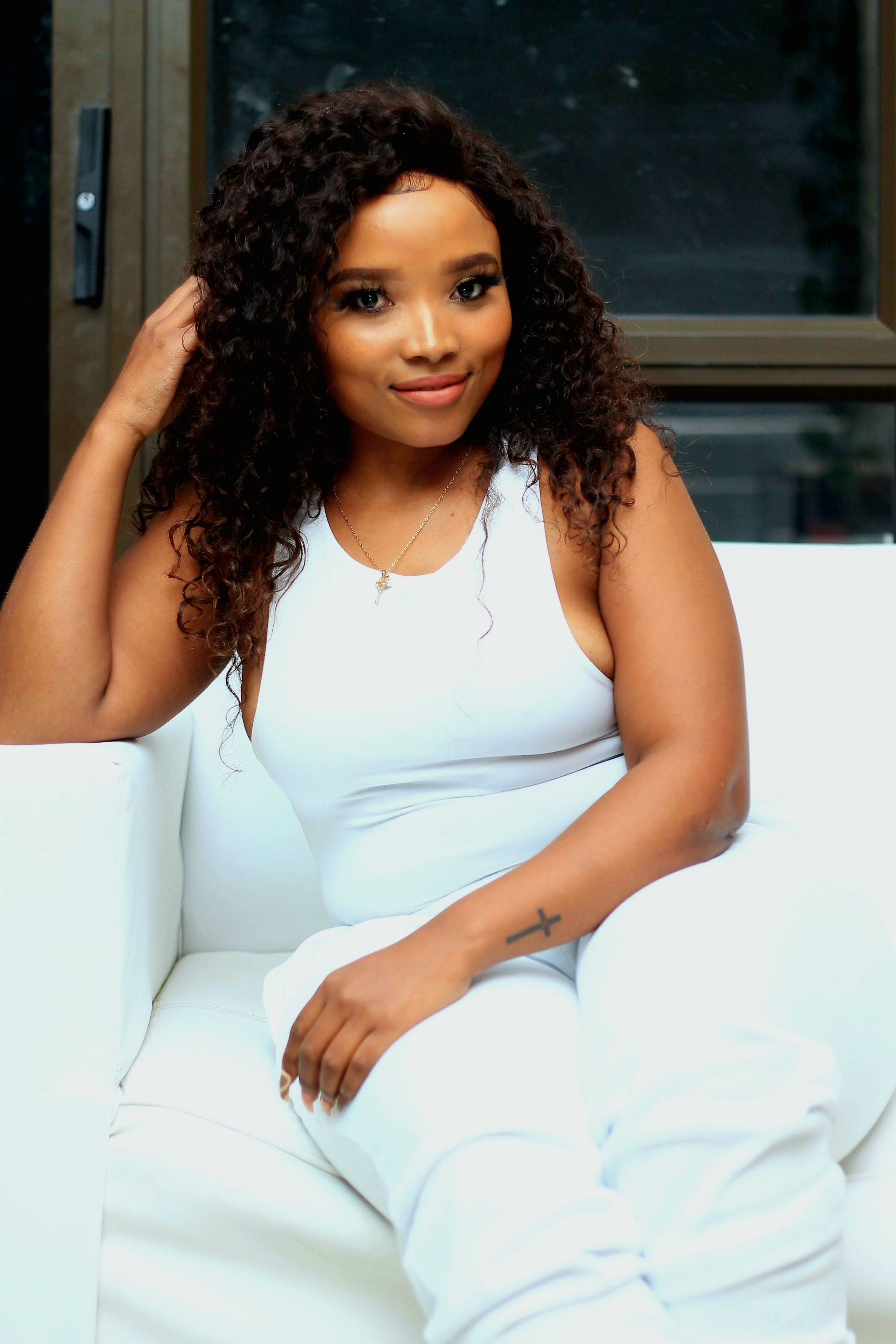 A young woman in casual attire sitting on a white sofa indoors, smiling gently.