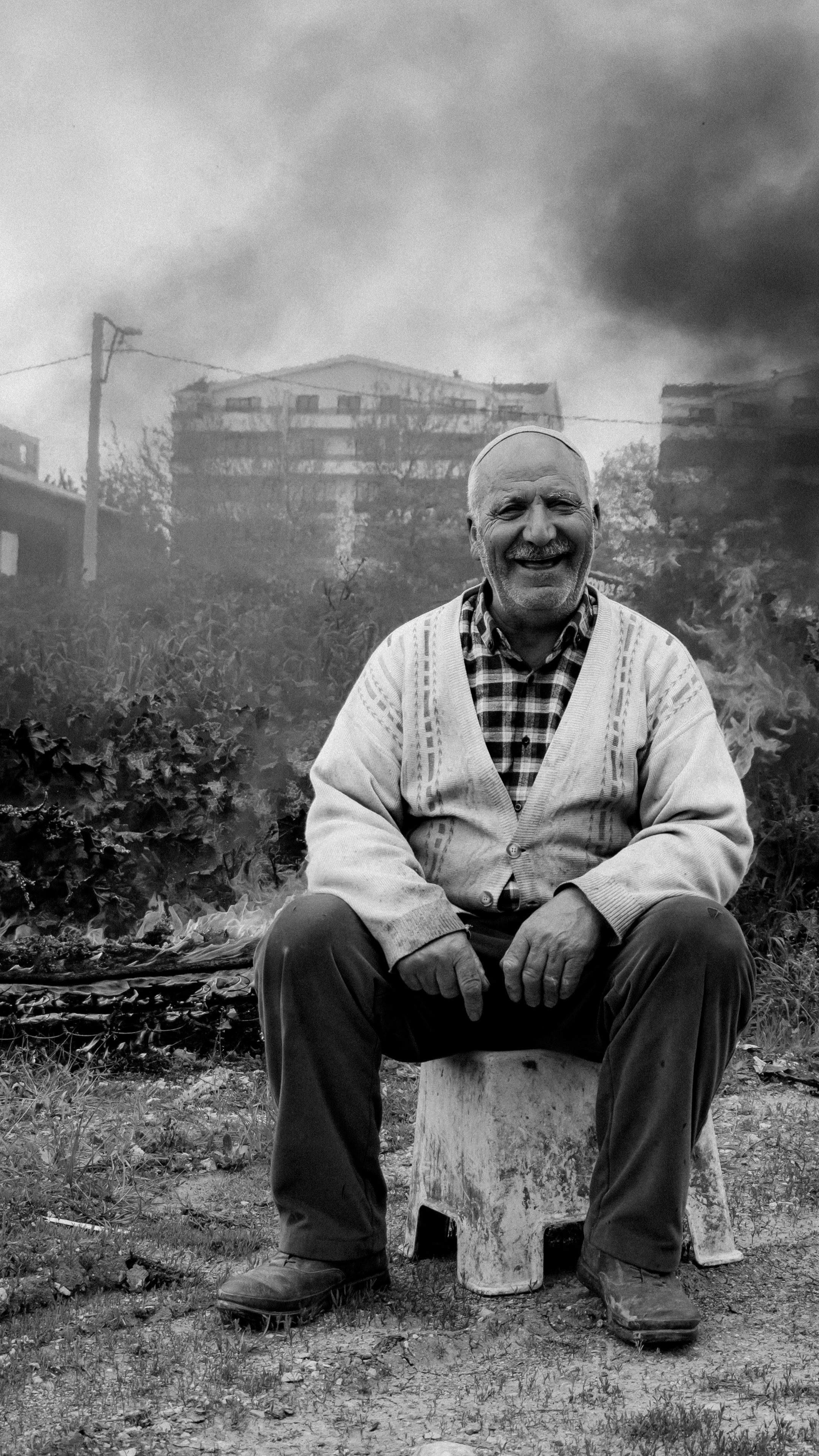 A senior man seated in an urban outdoor setting with buildings and smoke in the background.
