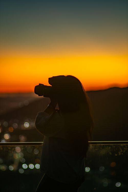 Free Silhouette of a photographer capturing a vibrant sunset with city lights in the background, creating a dramatic scene. Stock Photo