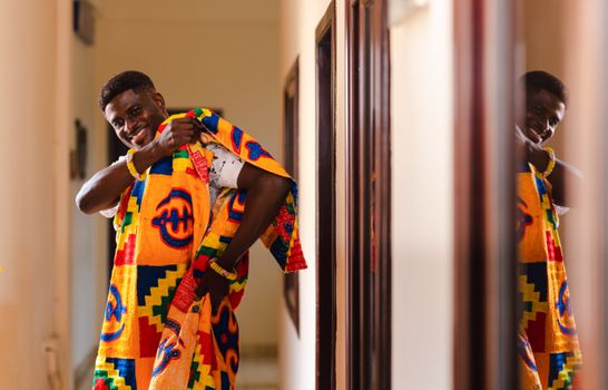A smiling man in vibrant traditional attire stands in a hallway with a mirrored reflection.