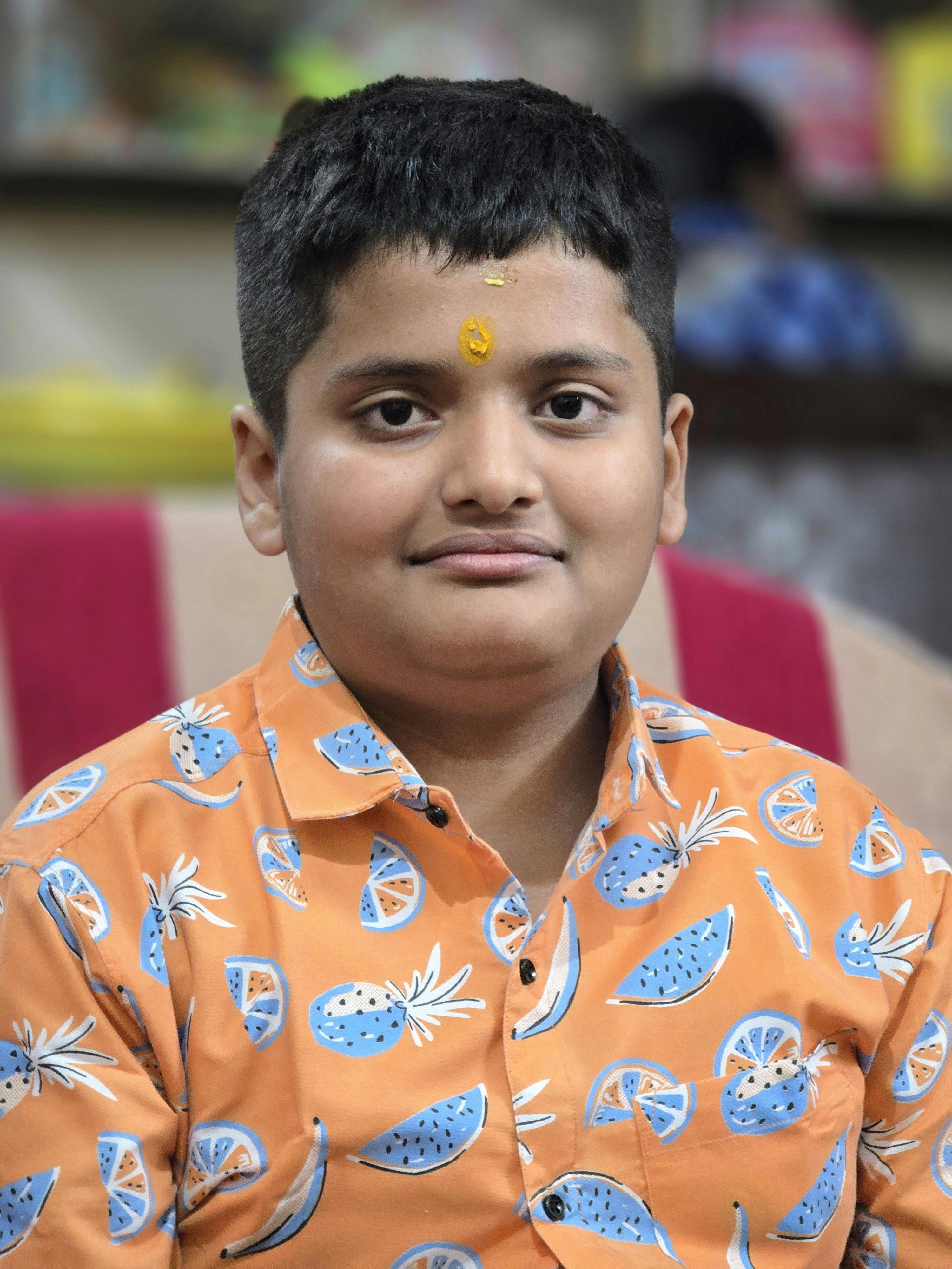 A young boy smiles indoors wearing a colorful patterned shirt, exuding warmth and charm.