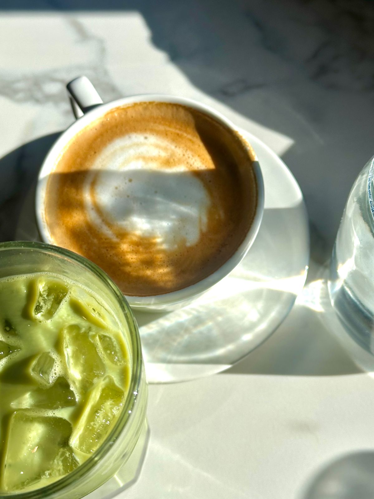 Iced matcha and a cup of coffee sharing a sunlit marble table