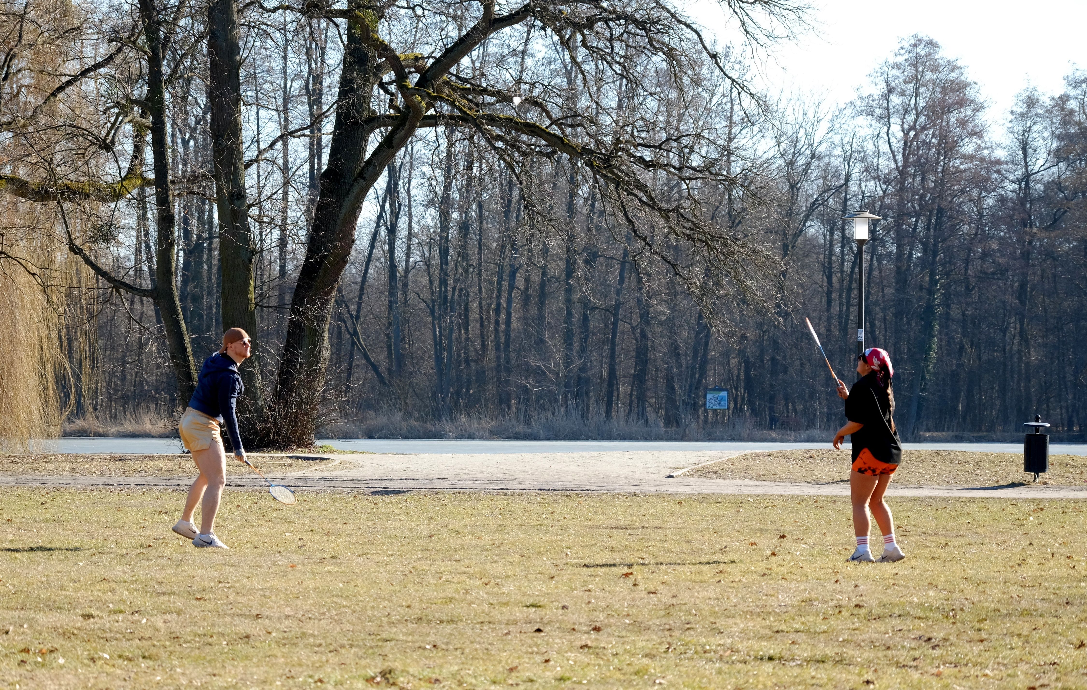 Two people playing badminton in a sunny park surrounded by bare trees.