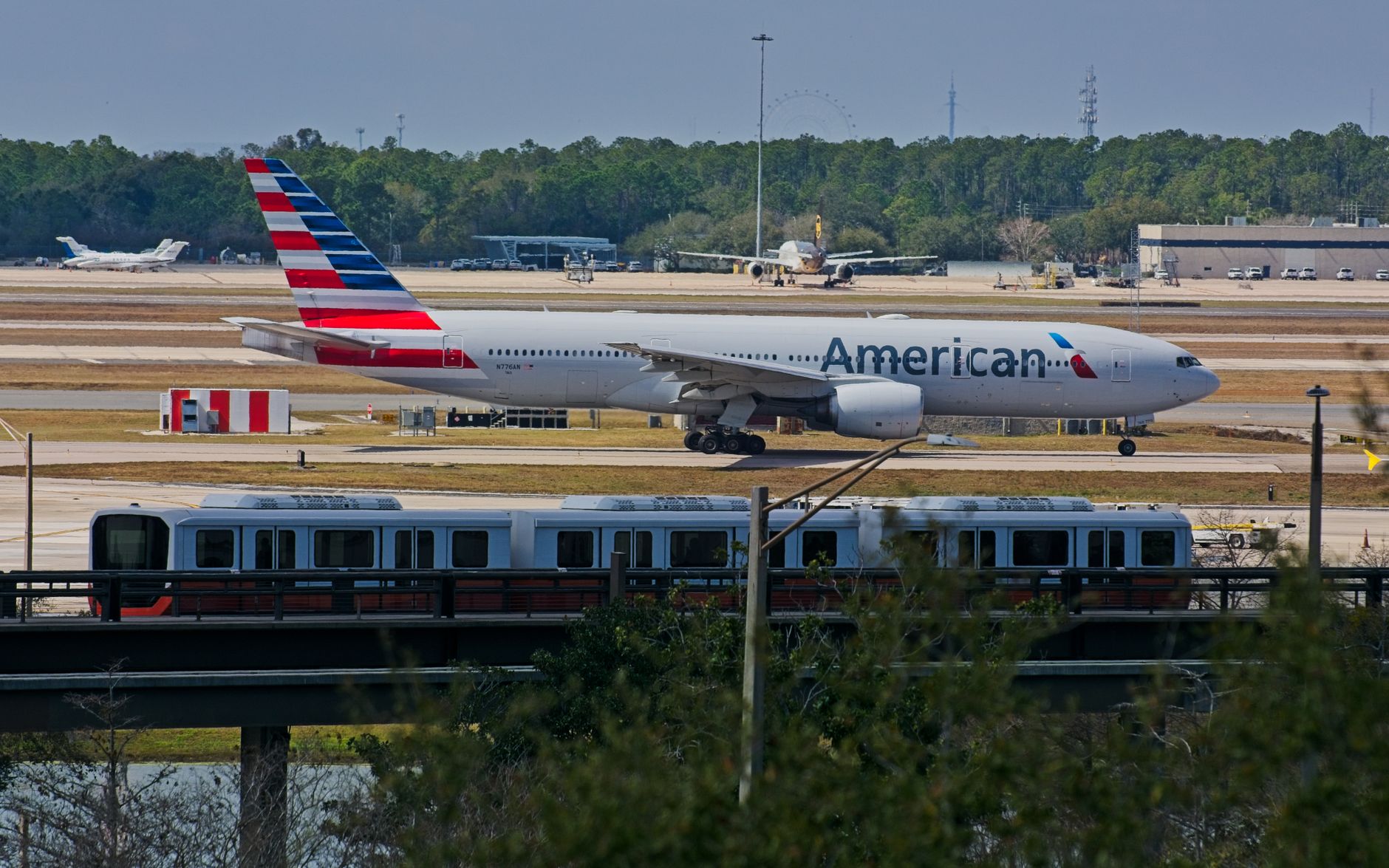 American Airlines Boeing 777 taxiing at Orlando International Airport with city rail in foreground.