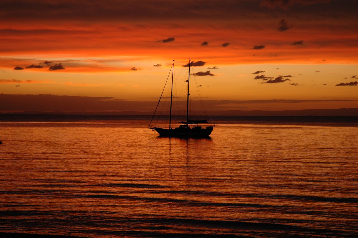 A sailboat at anchor, silhouetted against a deep orange and red sunset over calm water, back in port after the day's diving is logged.