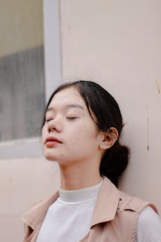 这是一幅特写肖像，画面平静，一位年轻的亚洲女子闭着眼睛，白天靠墙而立