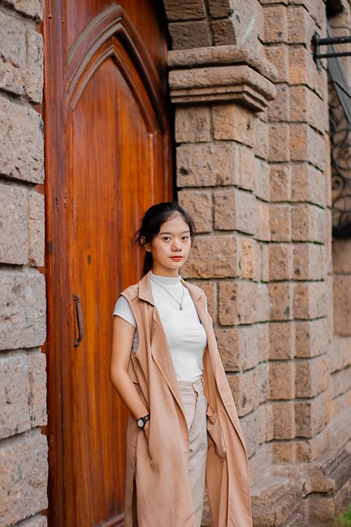 Free Calm Asian female teenager with dark hair in stylish outfit standing near aged stone building with wooden door with hands in pockets and looking at camera Stock Photo