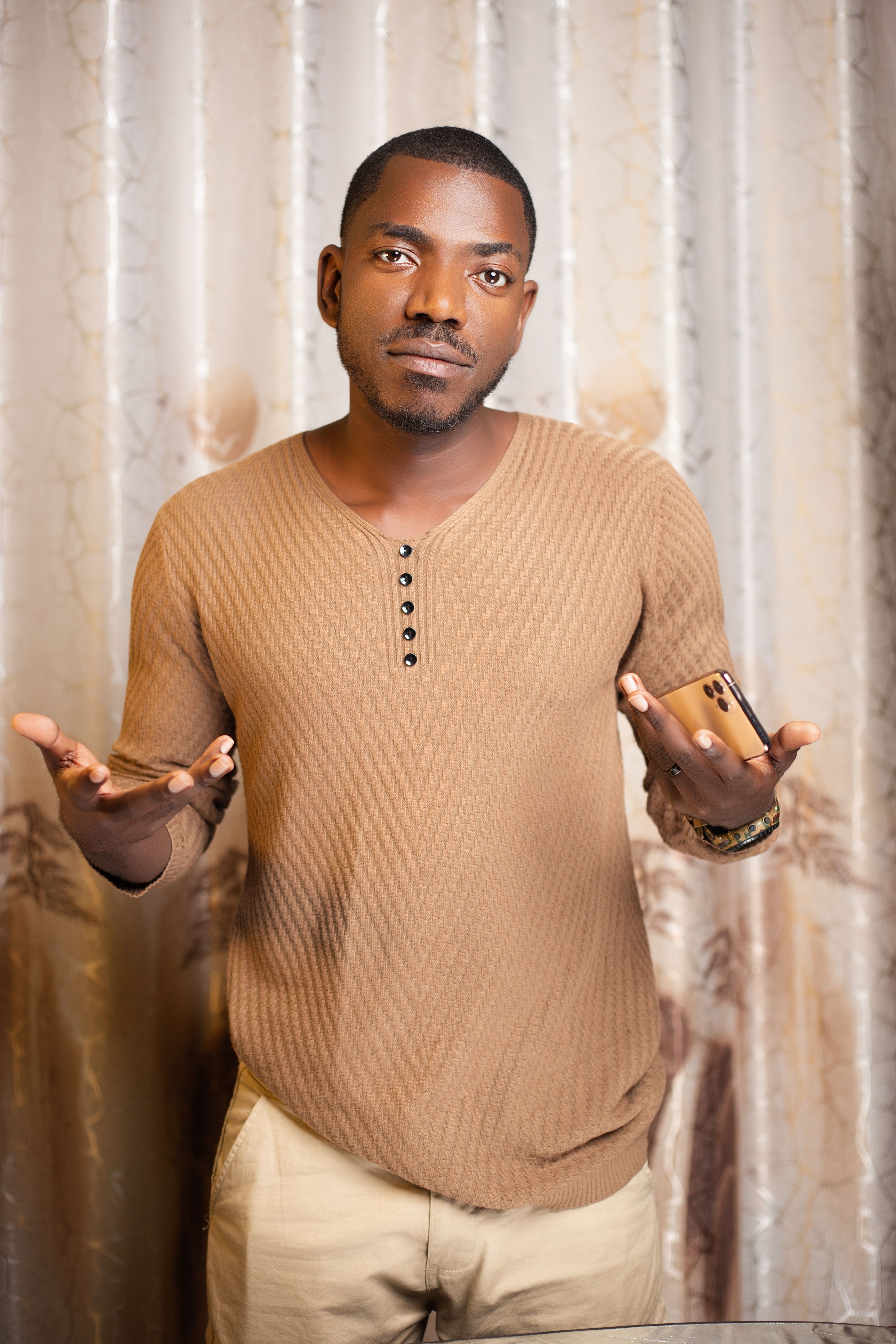 A young man in casual attire gestures with his hands while standing indoors against a decorative curtain.