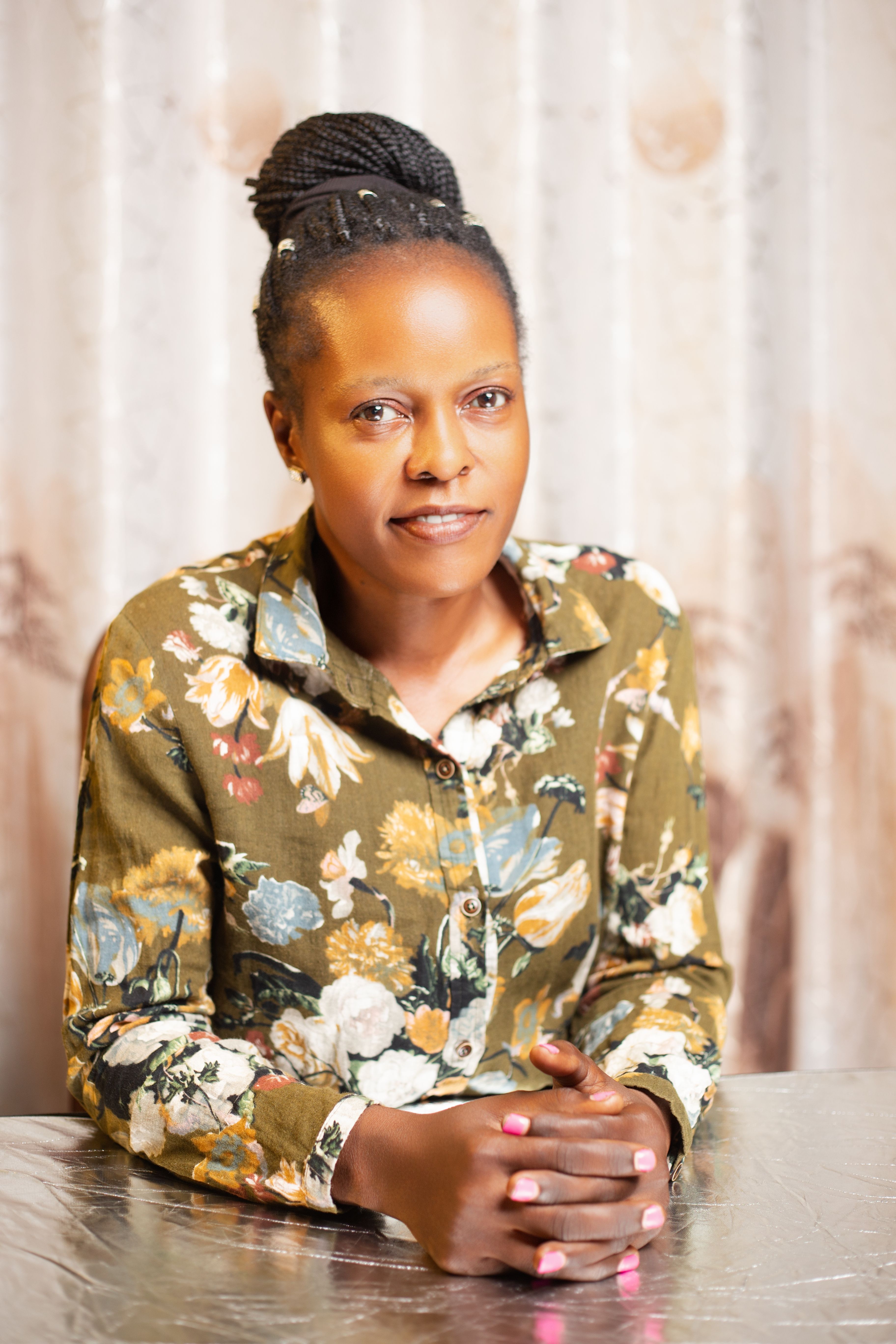 A woman in a floral shirt poses indoors against a beige curtain backdrop.