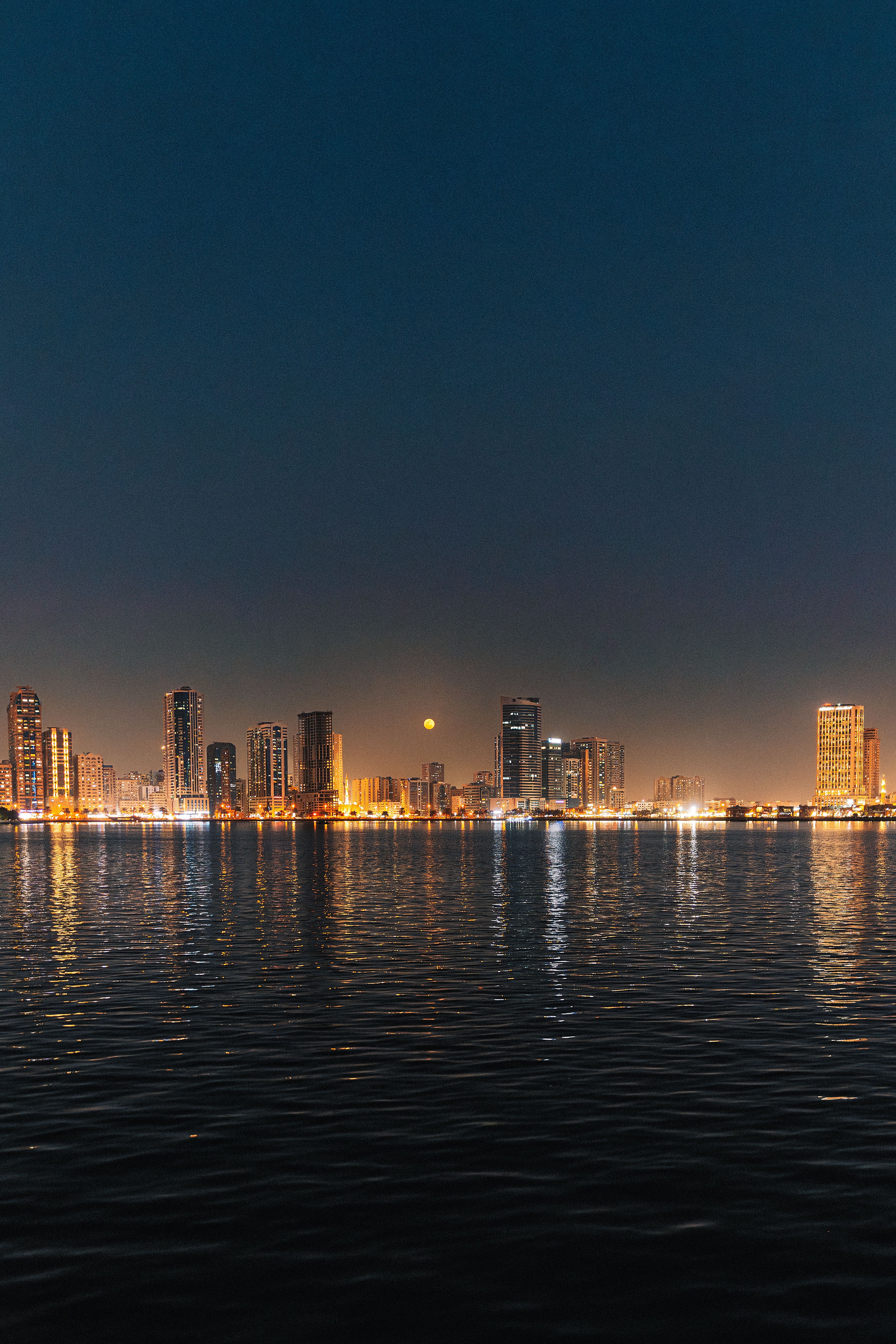 Stunning night view of a city skyline illuminated under a moonlit sky, reflected on a calm body of water.