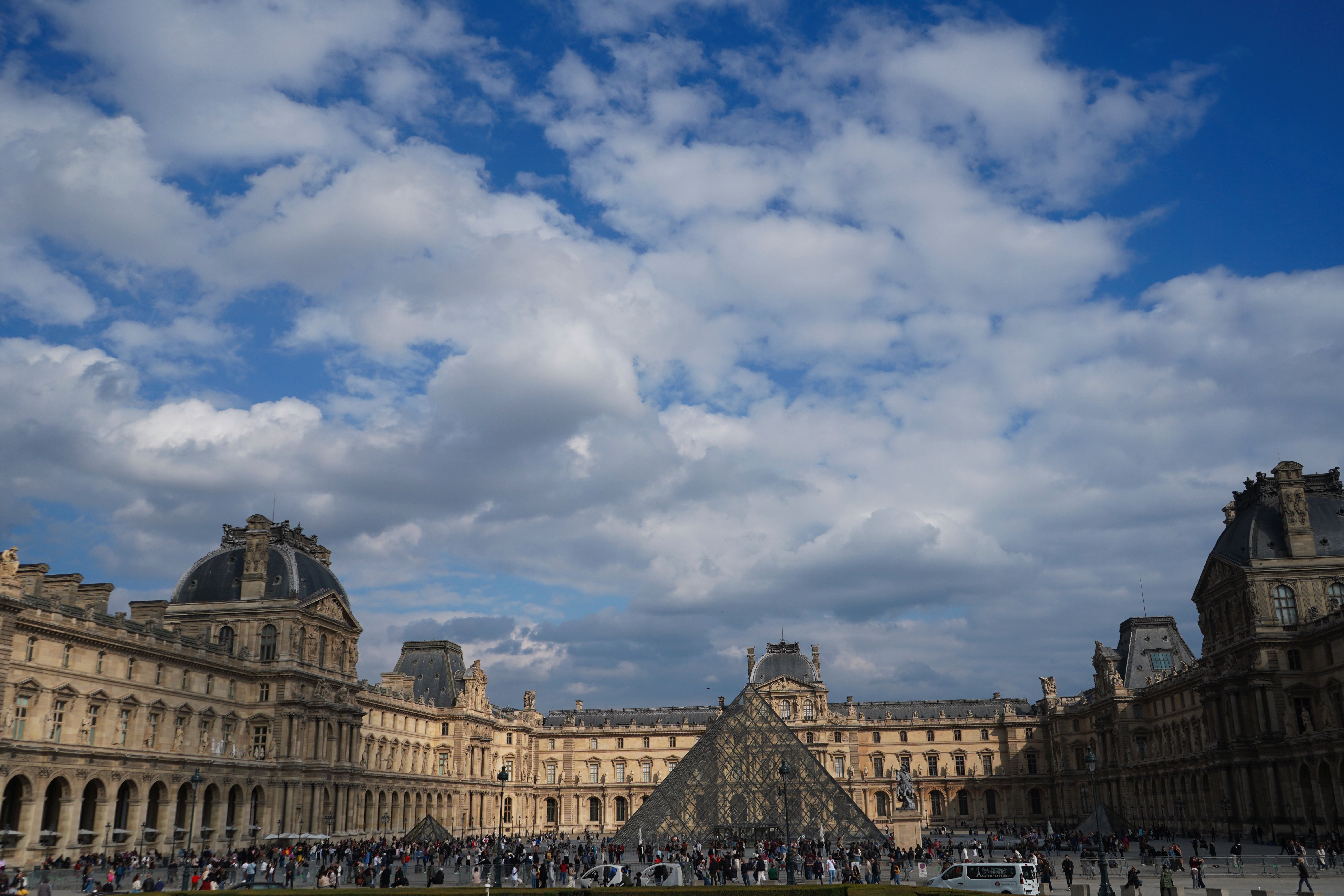 Louvre Museum courtyard in Paris with iconic glass pyramid under a blue sky, crowded with visitors.
