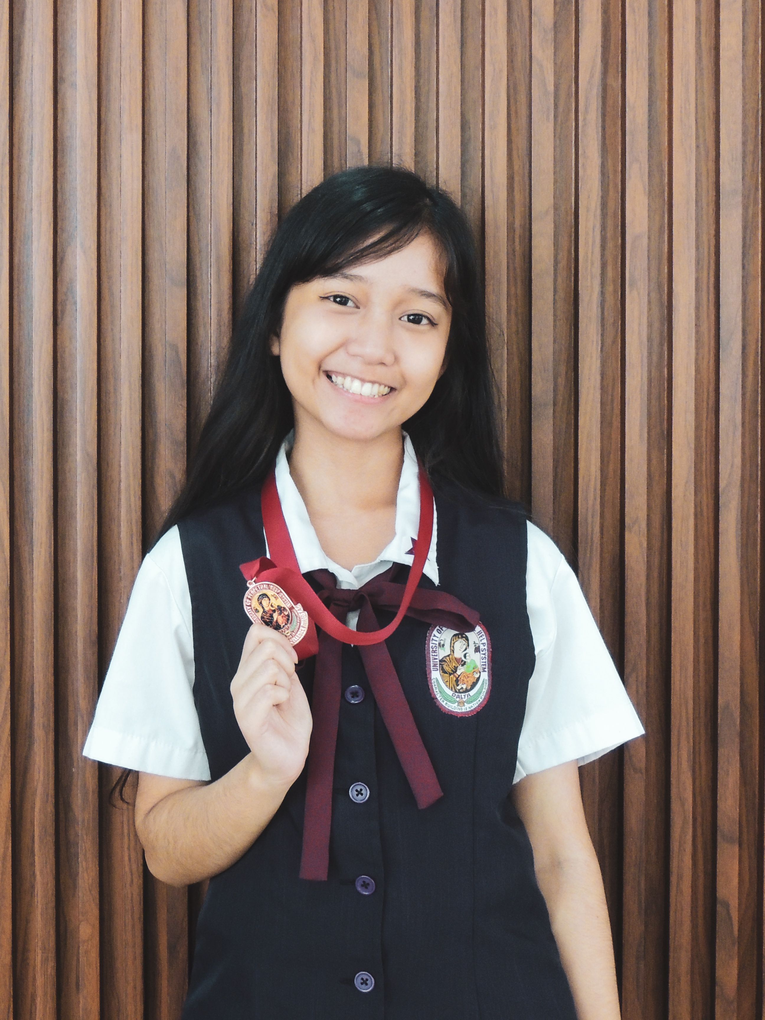 Smiling student in uniform holding a medal against a wooden backdrop.