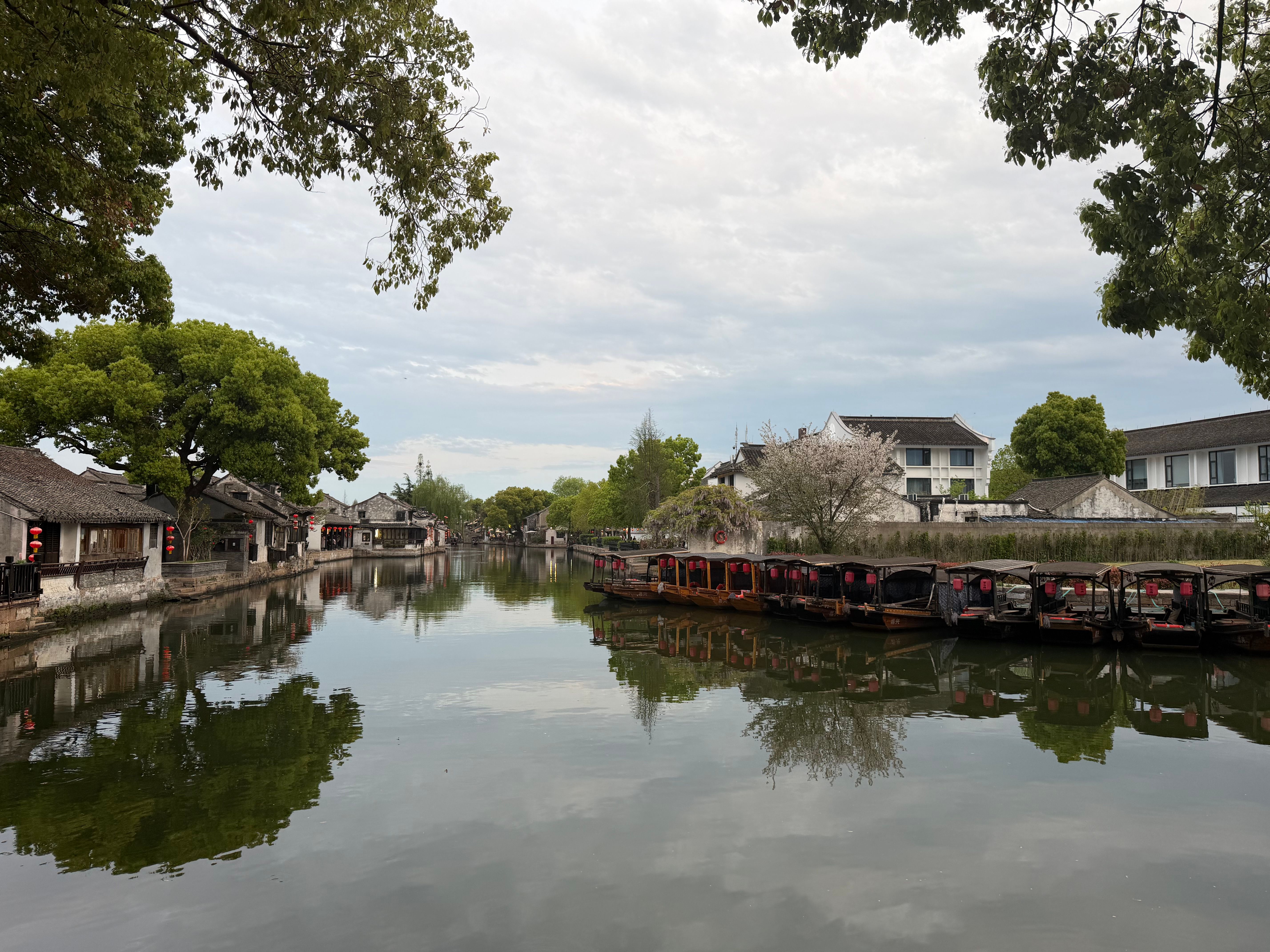 Tranquil view of a historic Chinese village with boats along a serene waterway under a cloudy sky.
