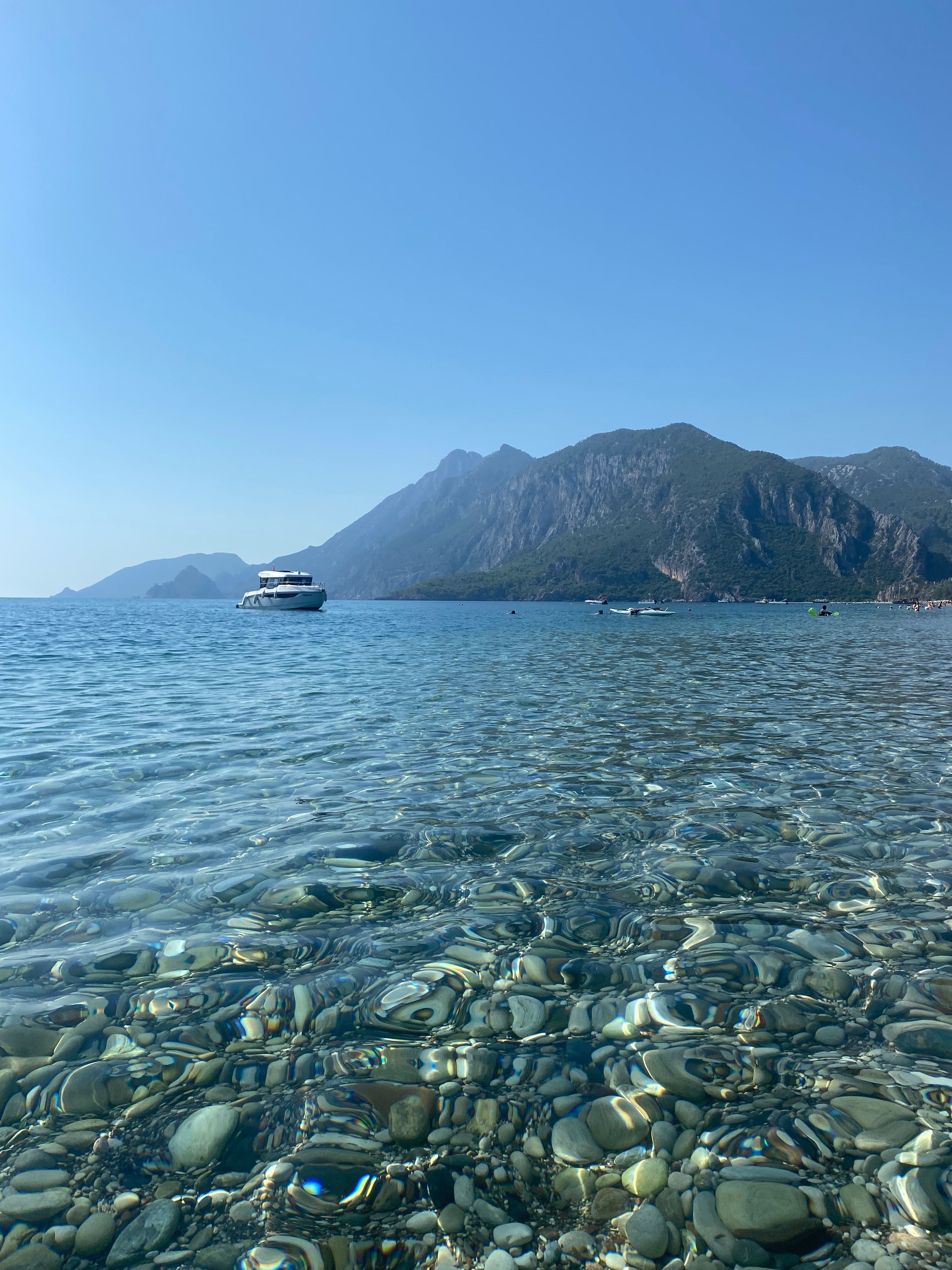 Clear waters and mountainous backdrop of a Mediterranean shore featuring a distant yacht.