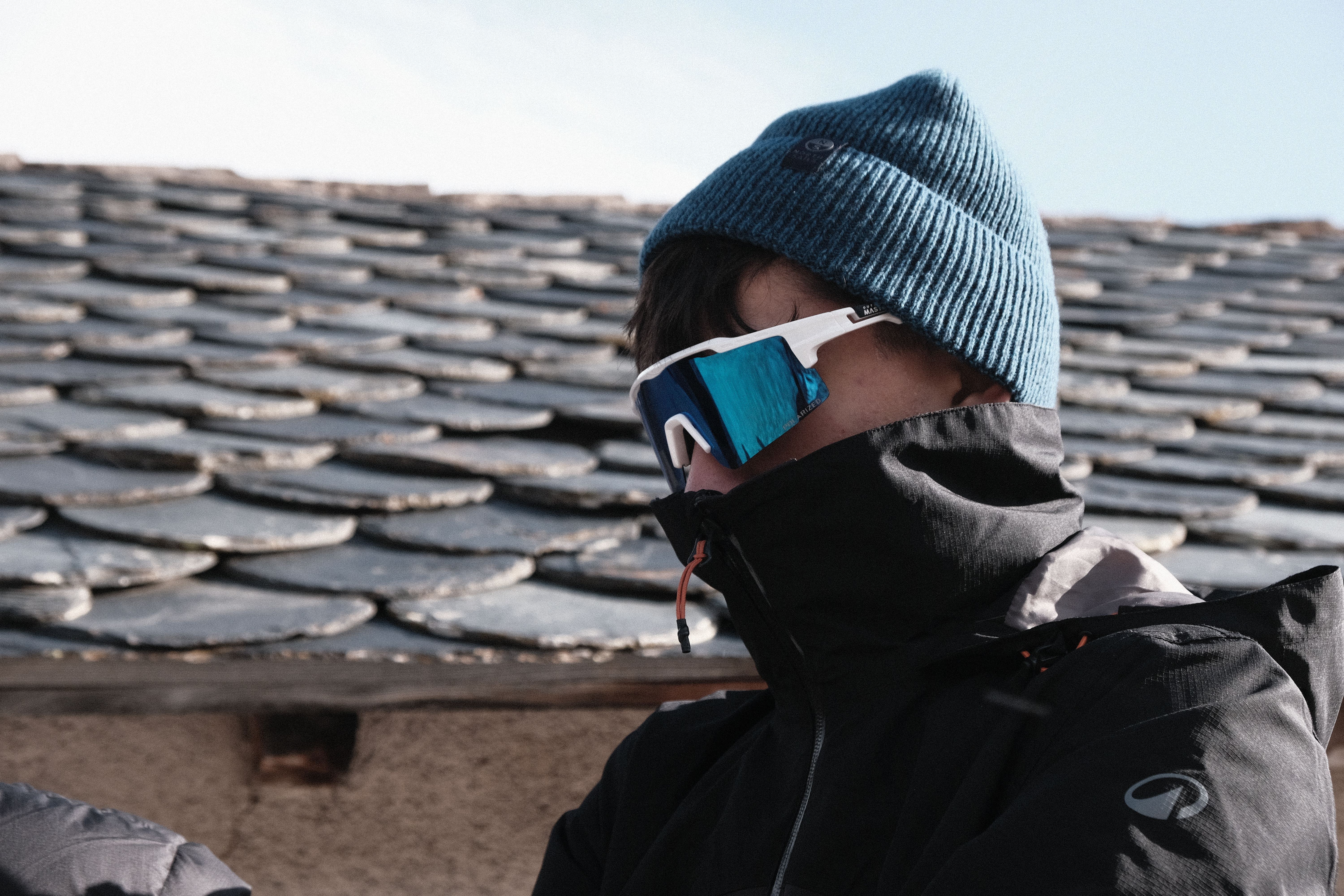 A person bundled in winter attire wearing reflective sunglasses against a rustic shingle roof.