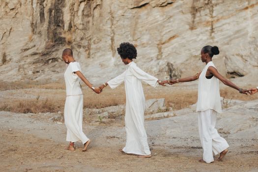Three women in white outfits holding hands and walking along a rocky outdoor cliffside, symbolizing friendship.