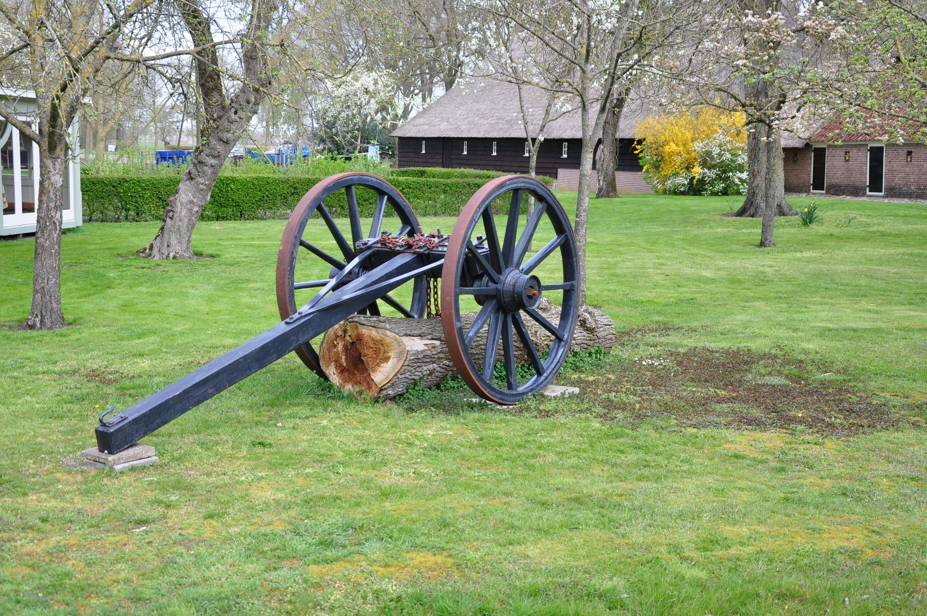 A vintage cannon rests on green grass with a backdrop of spring trees and rustic buildings.