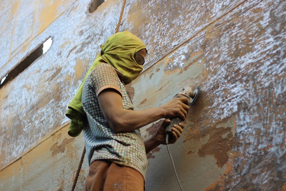 A worker in protective gear grinding a large metal surface at a shipyard.