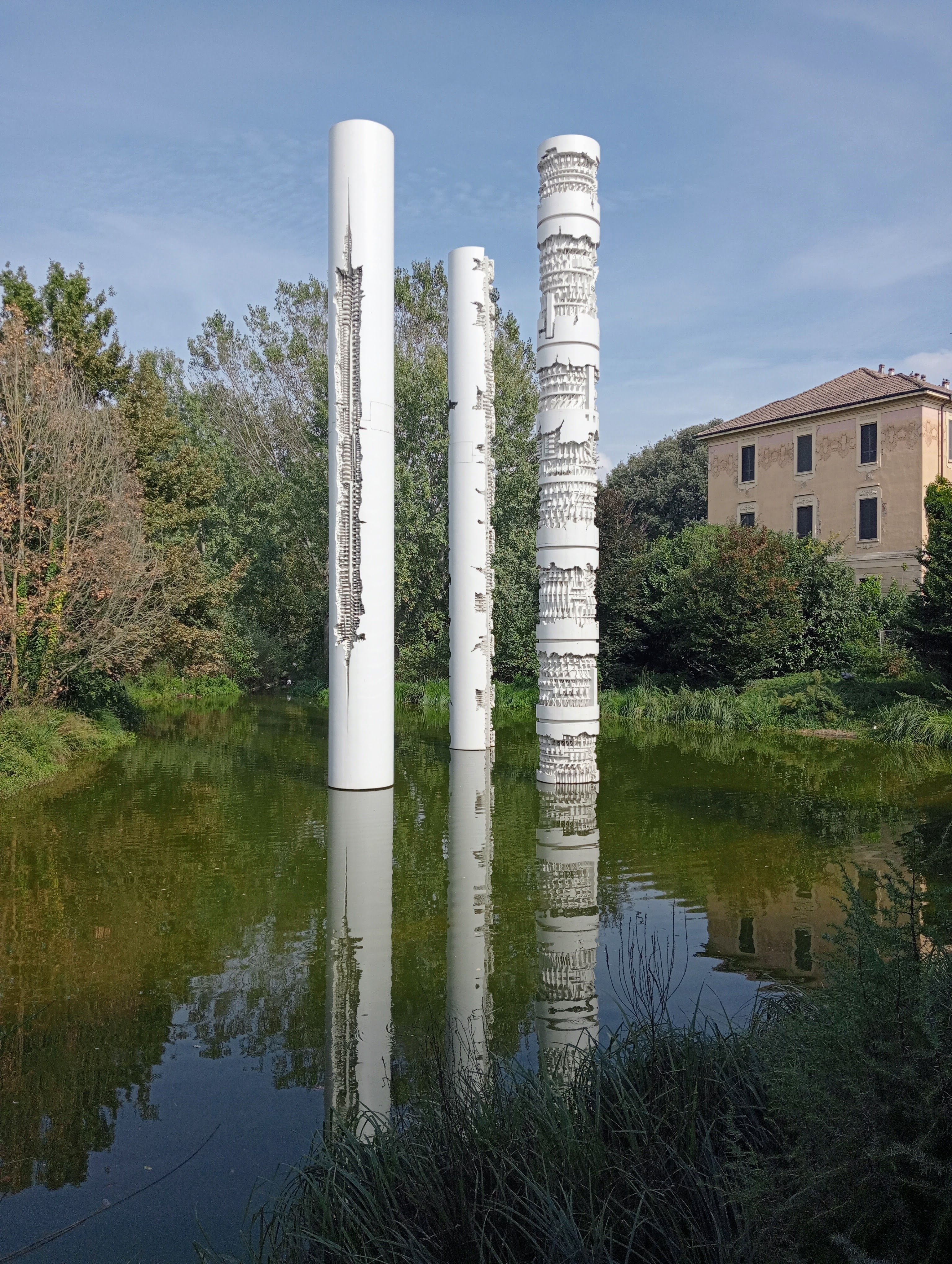 Sculpture poles reflecting in a serene pond with a villa backdrop in Pavia.
