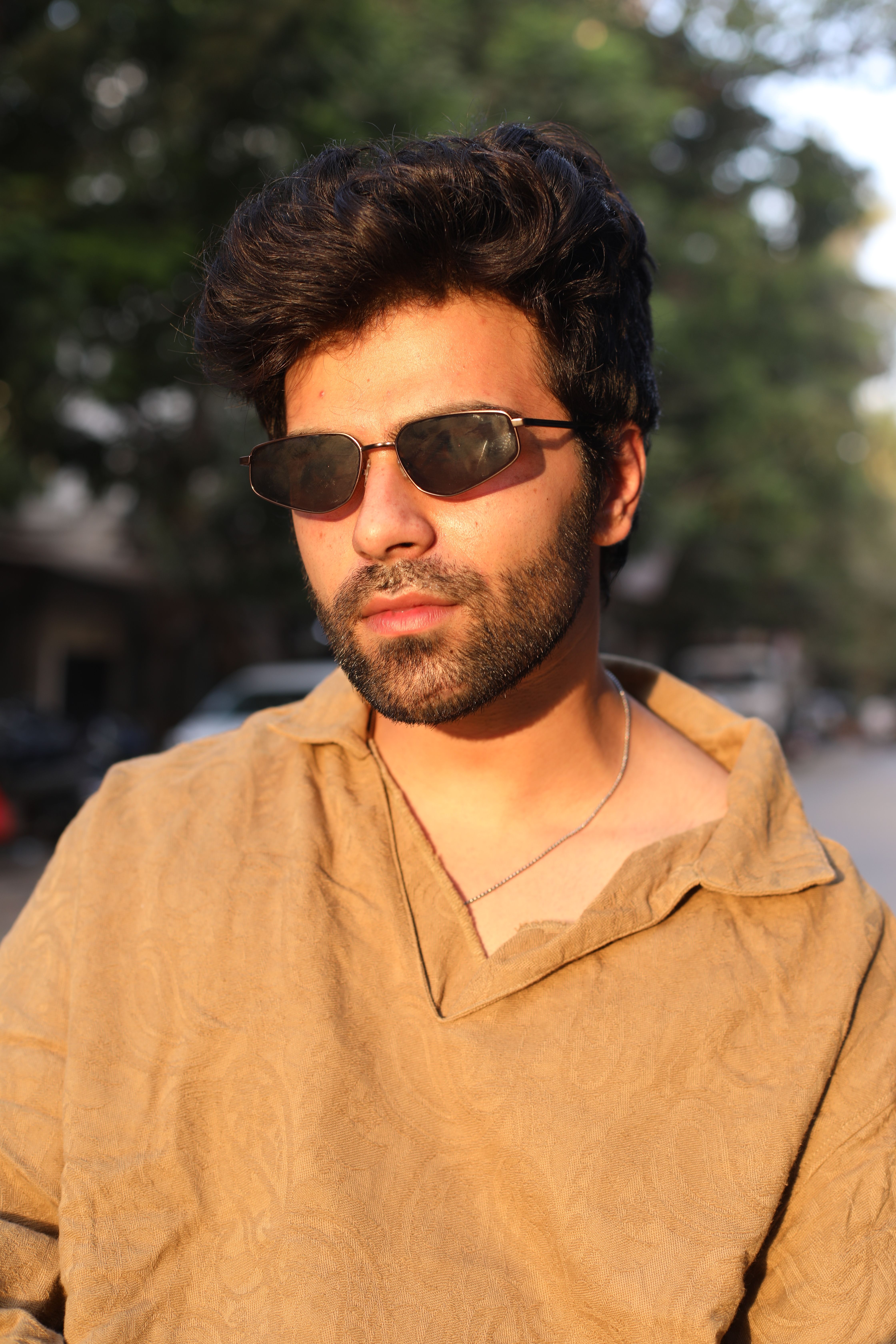 Stylish man in sunglasses and brown shirt posing outdoors during sunset.