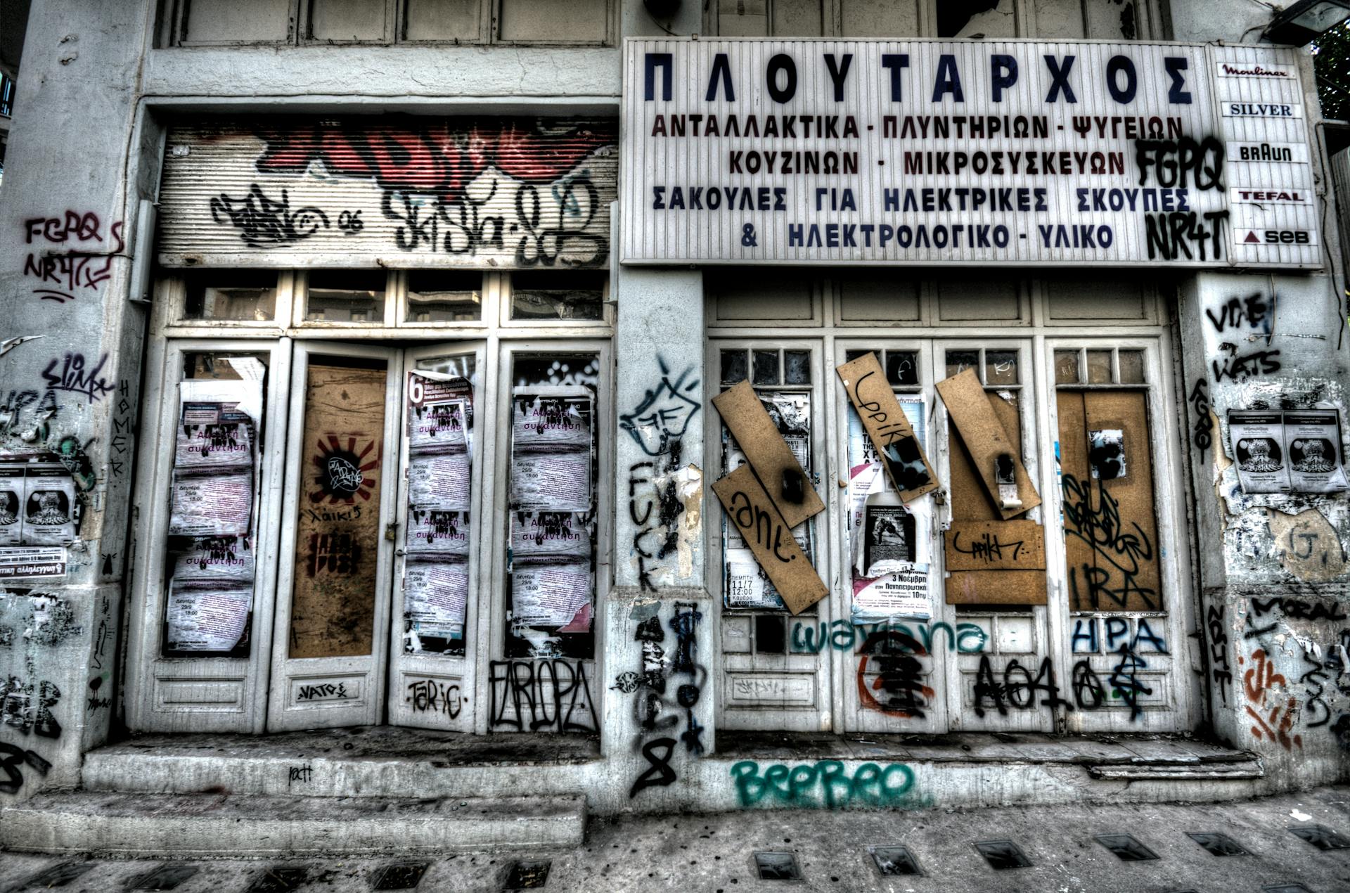 Abandoned graffiti-covered storefront featuring Greek signs and closed doors.