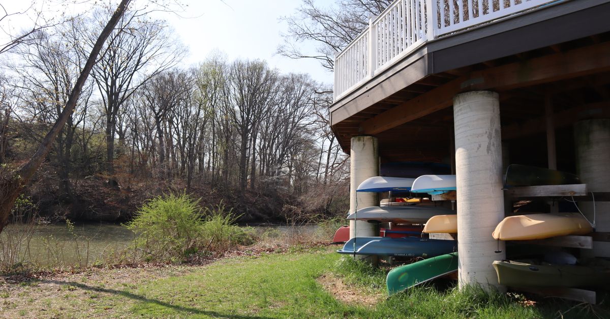A collection of kayaks under a deck beside a calm river surrounded by bare trees.