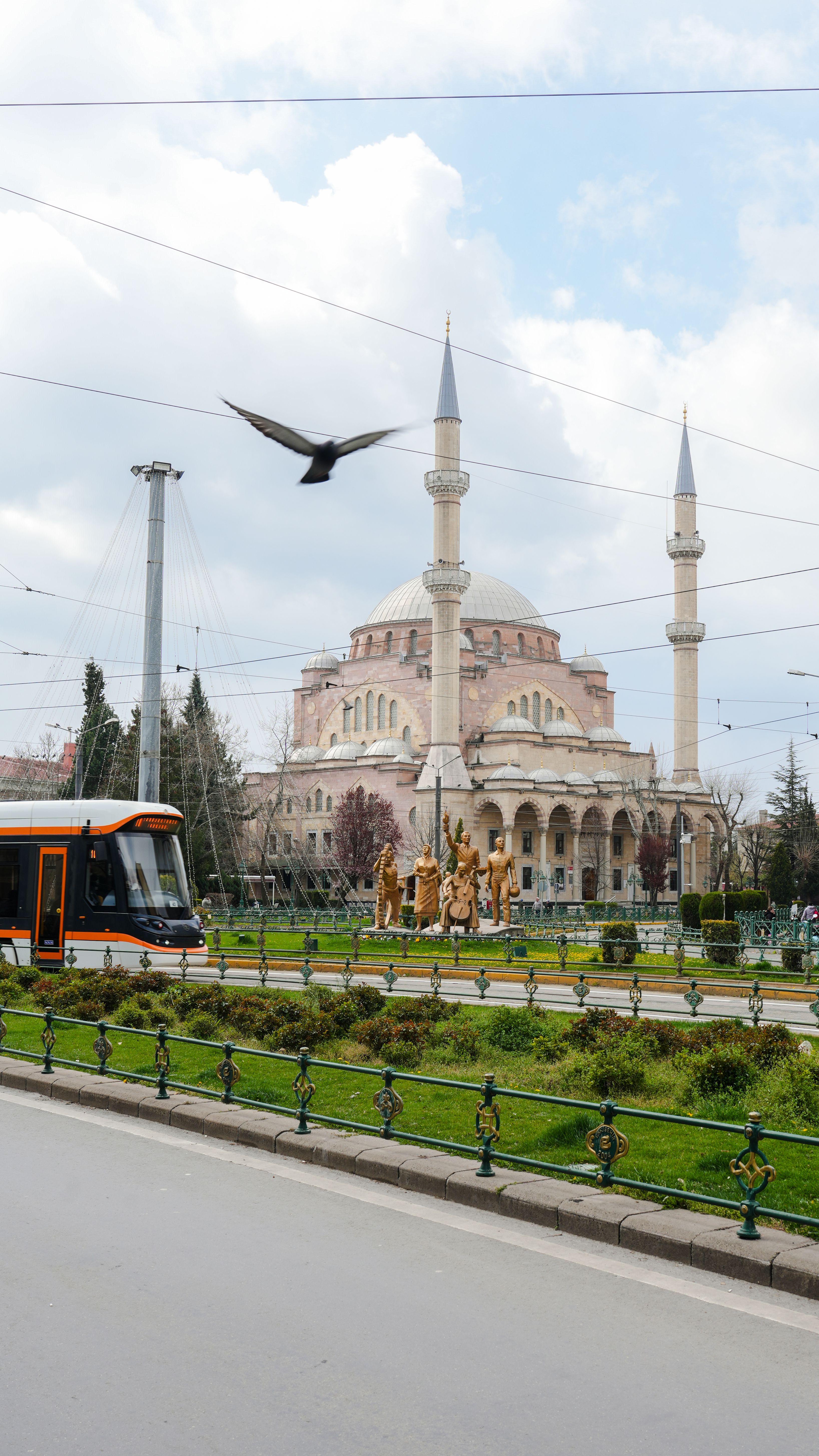 A tram moves past the historic architecture of a mosque in Istanbul with a bird flying overhead.