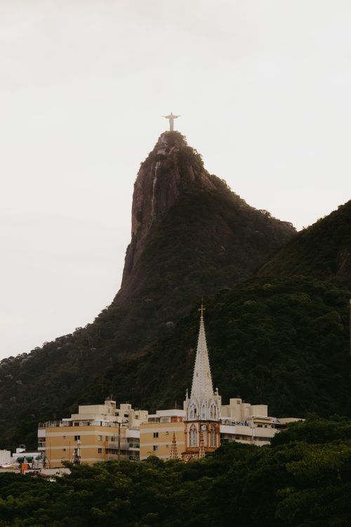 Free View of Christ the Redeemer statue above Rio de Janeiro, Brazil, with a church and lush greenery. Stock Photo