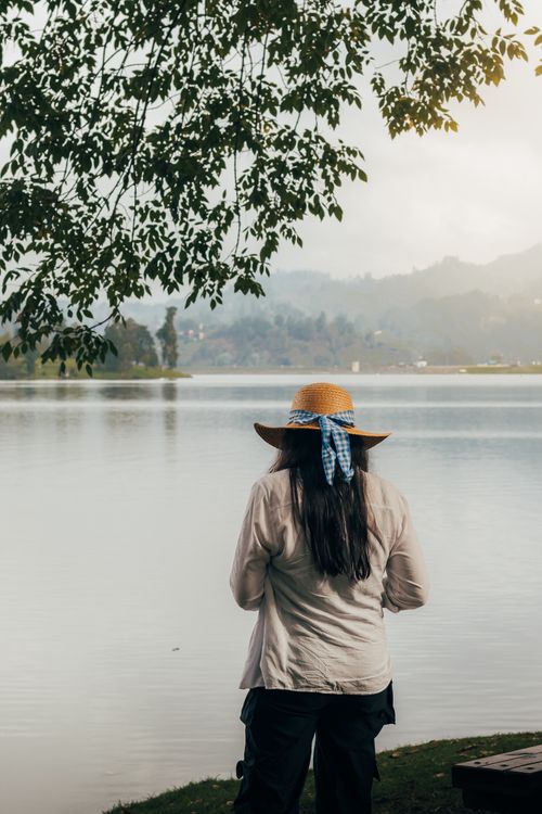 Free A woman wearing a straw hat gazes at a serene lake with mountains in the background. Stock Photo