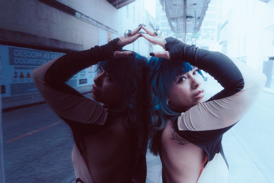 Artistic portrait of a stylish woman with blue hair posing against a reflective mirror on an urban street.