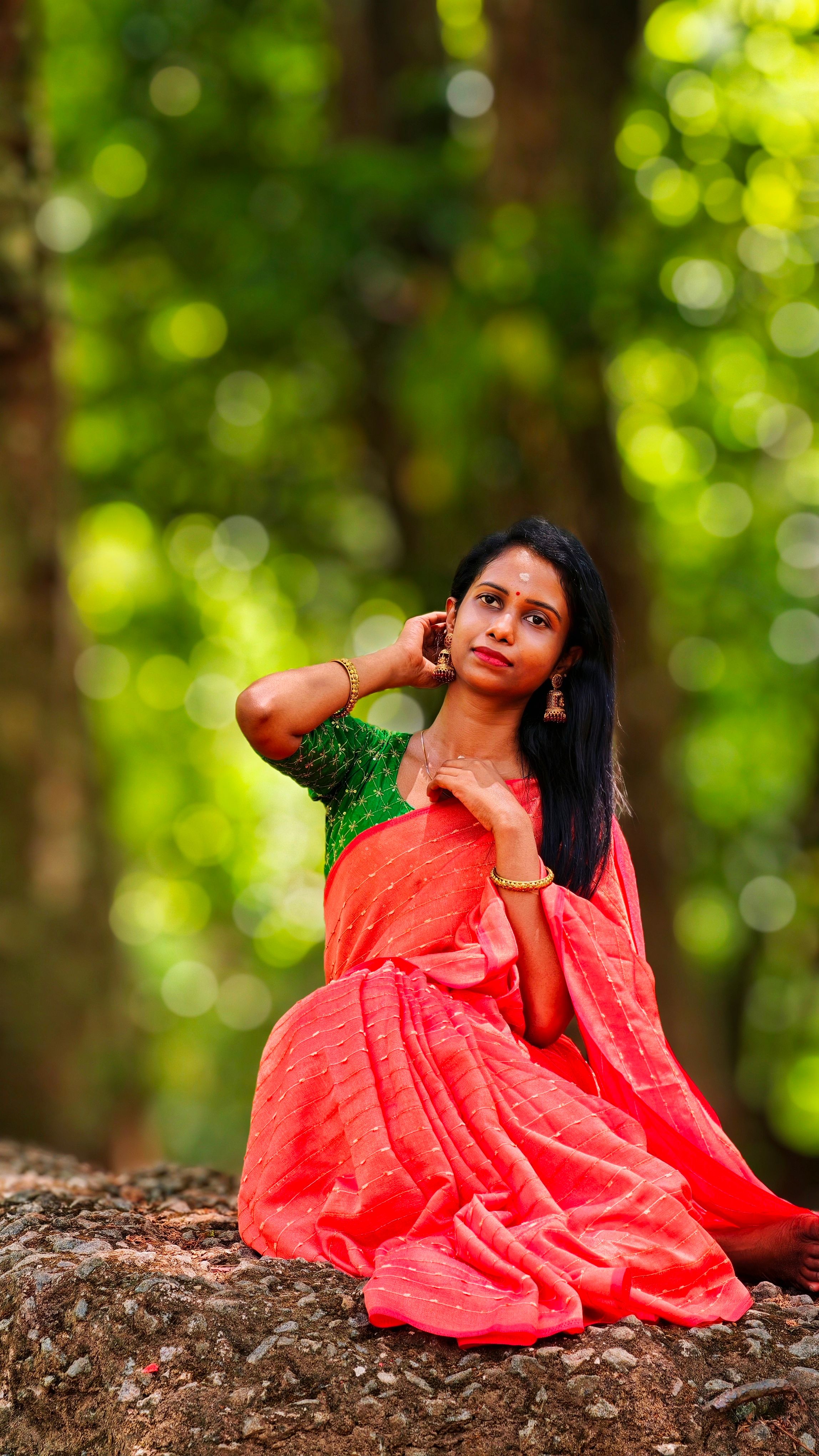Portrait of an elegant Indian woman wearing a red saree in an outdoor forest setting, exuding graceful beauty.