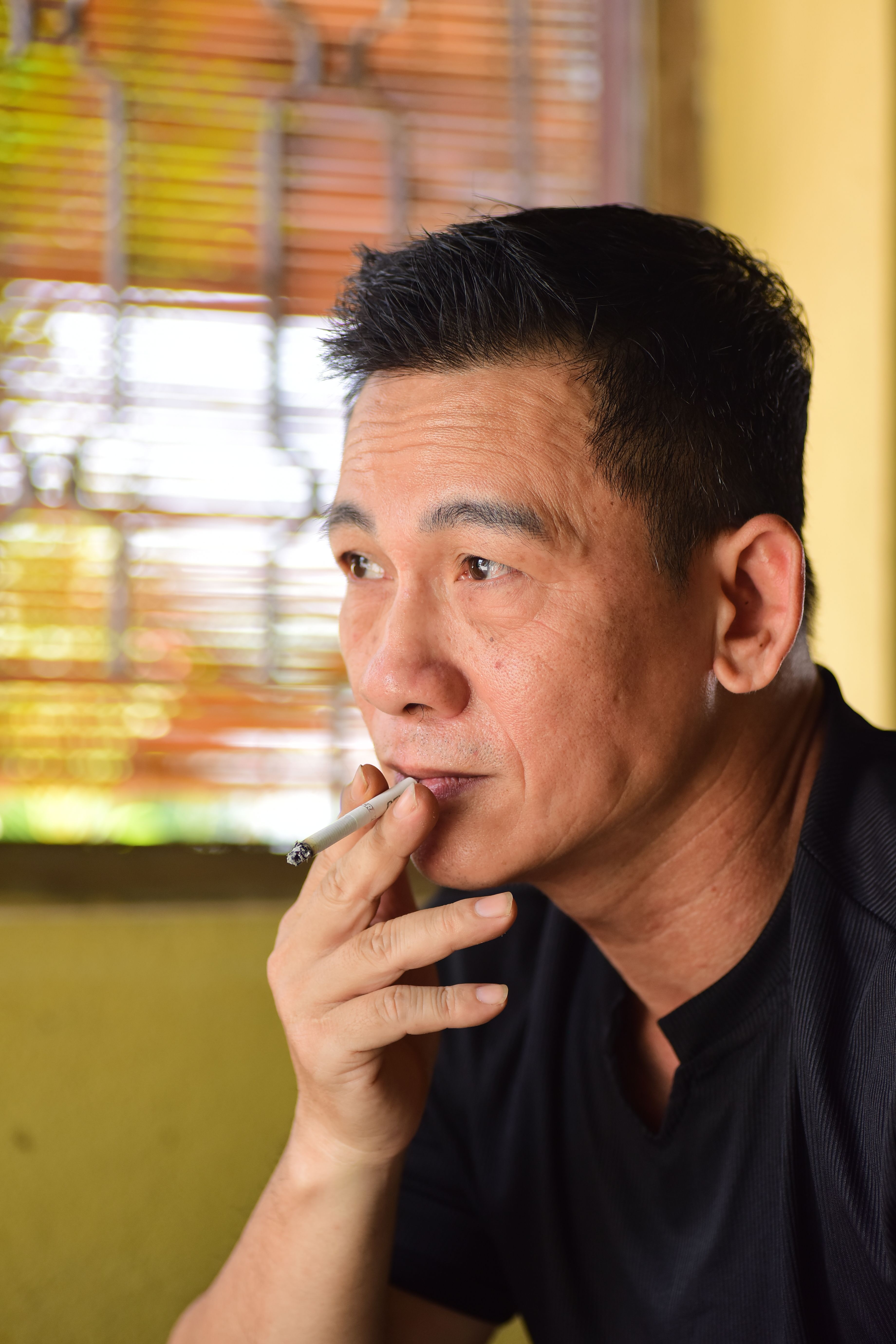 Thoughtful adult man seated indoors holding a cigarette, gazing into distance.