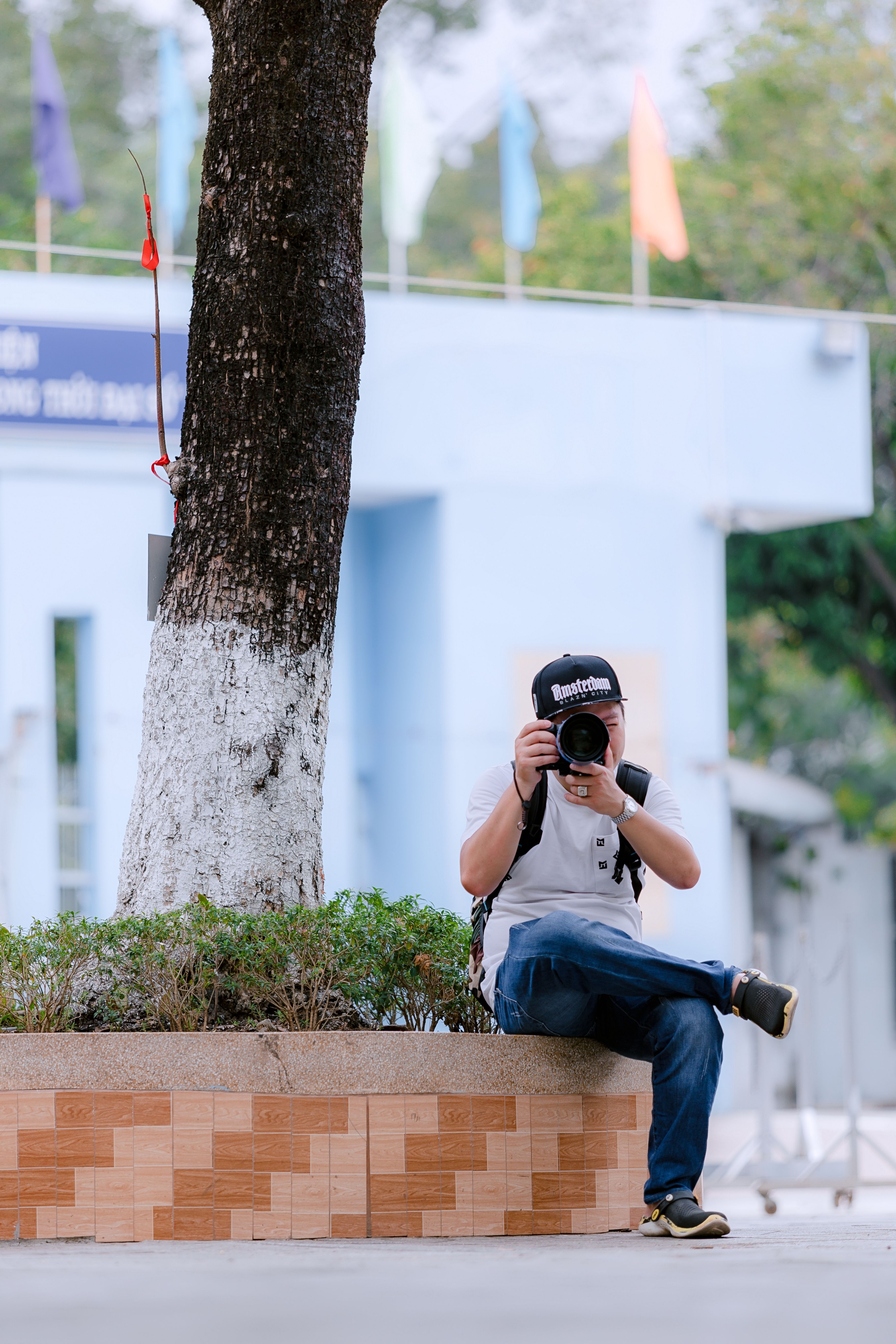 A man with a camera sits under a tree, capturing the surroundings on a sunny day.