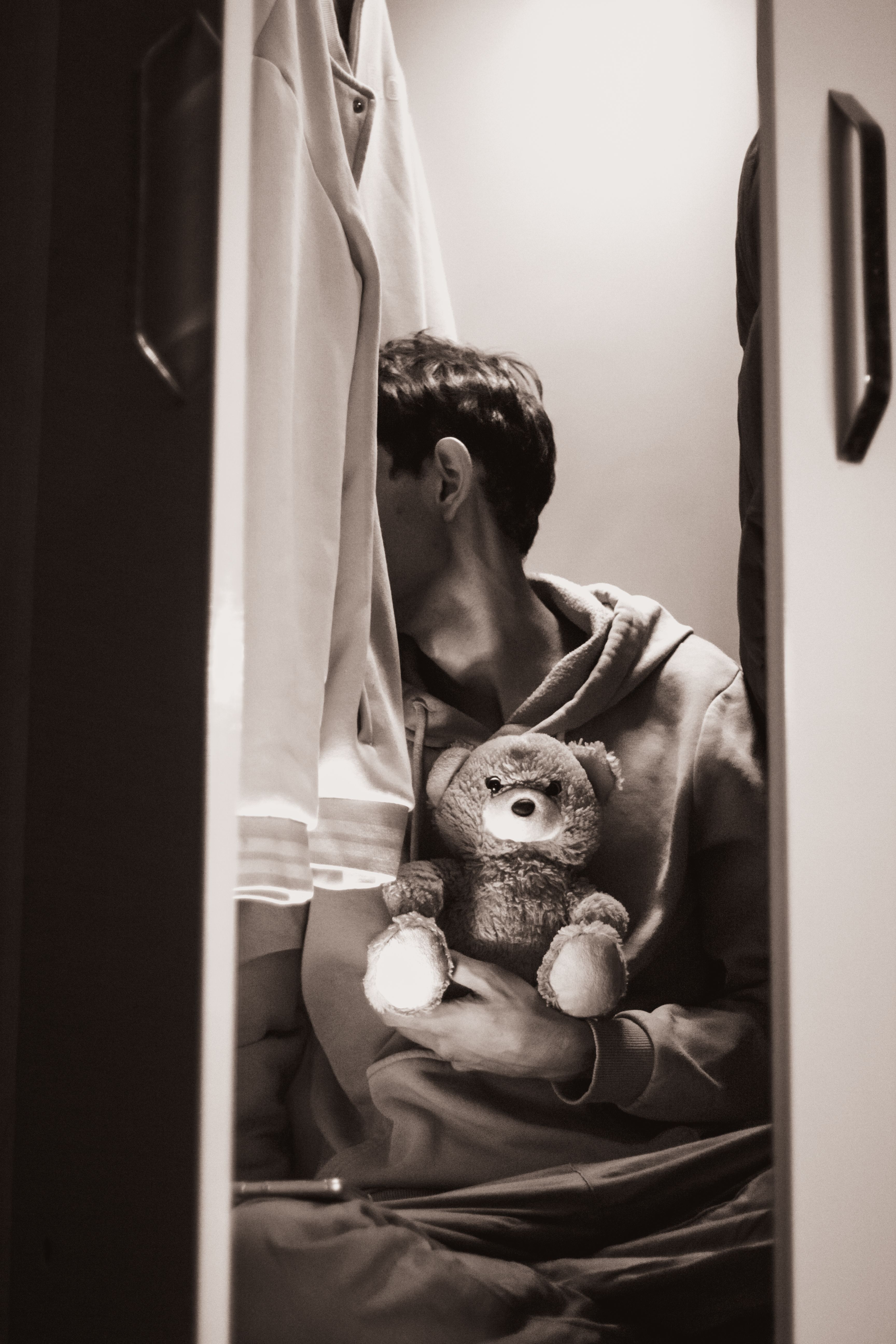 Fotomural Una persona está sentada en un armario con poca luz, sosteniendo un osito de peluche, transmitiendo una sensación de soledad.