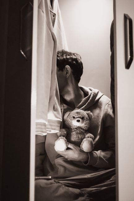 Fotomural Una persona está sentada en un armario con poca luz, sosteniendo un osito de peluche, transmitiendo una sensación de soledad.