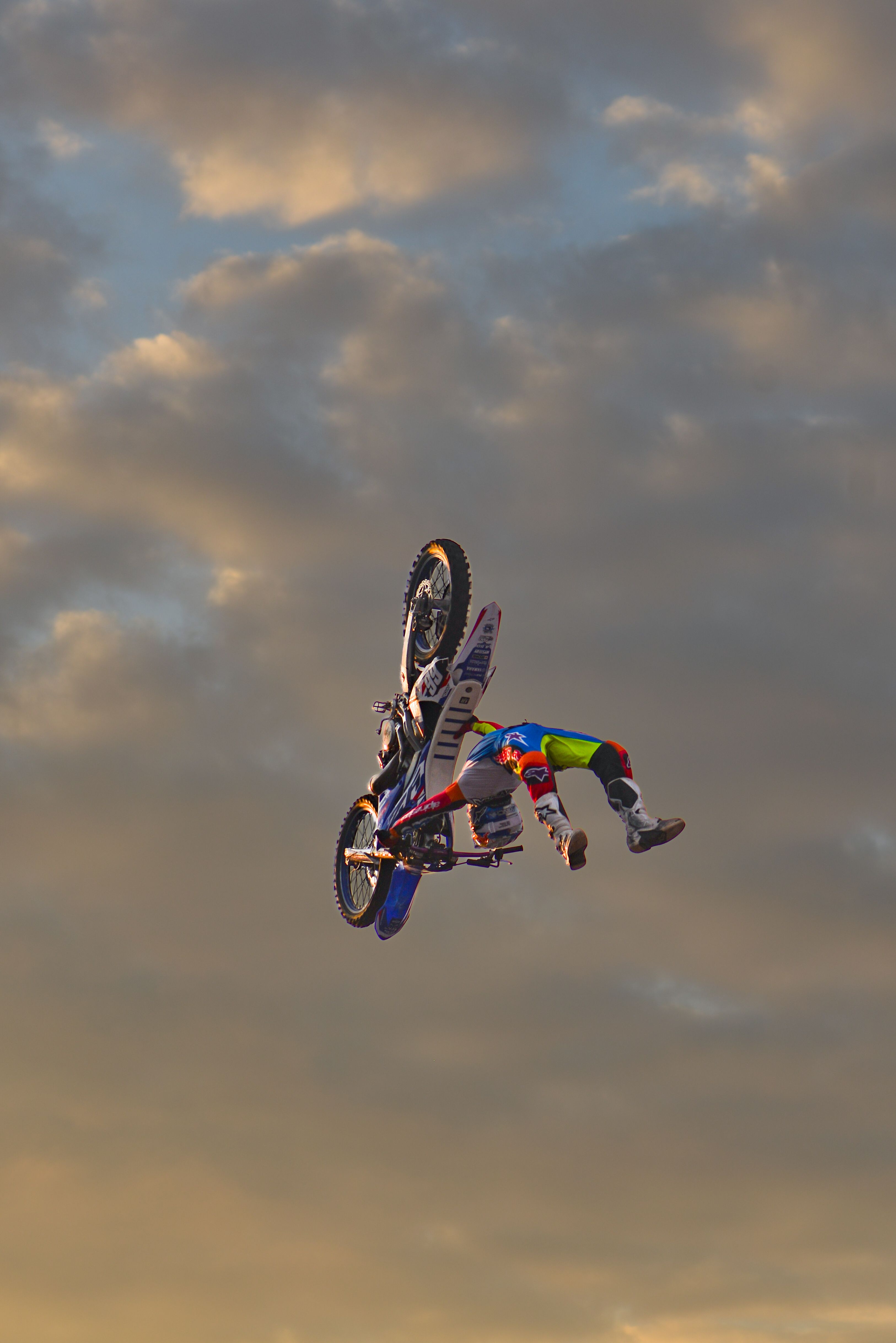 A motorcyclist in vibrant gear performs an aerial stunt against a dramatic sky at twilight.