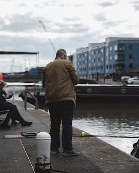 在港口码头上，一名男子正悠闲地钓着鱼，四周环绕着城市建筑，天空多云