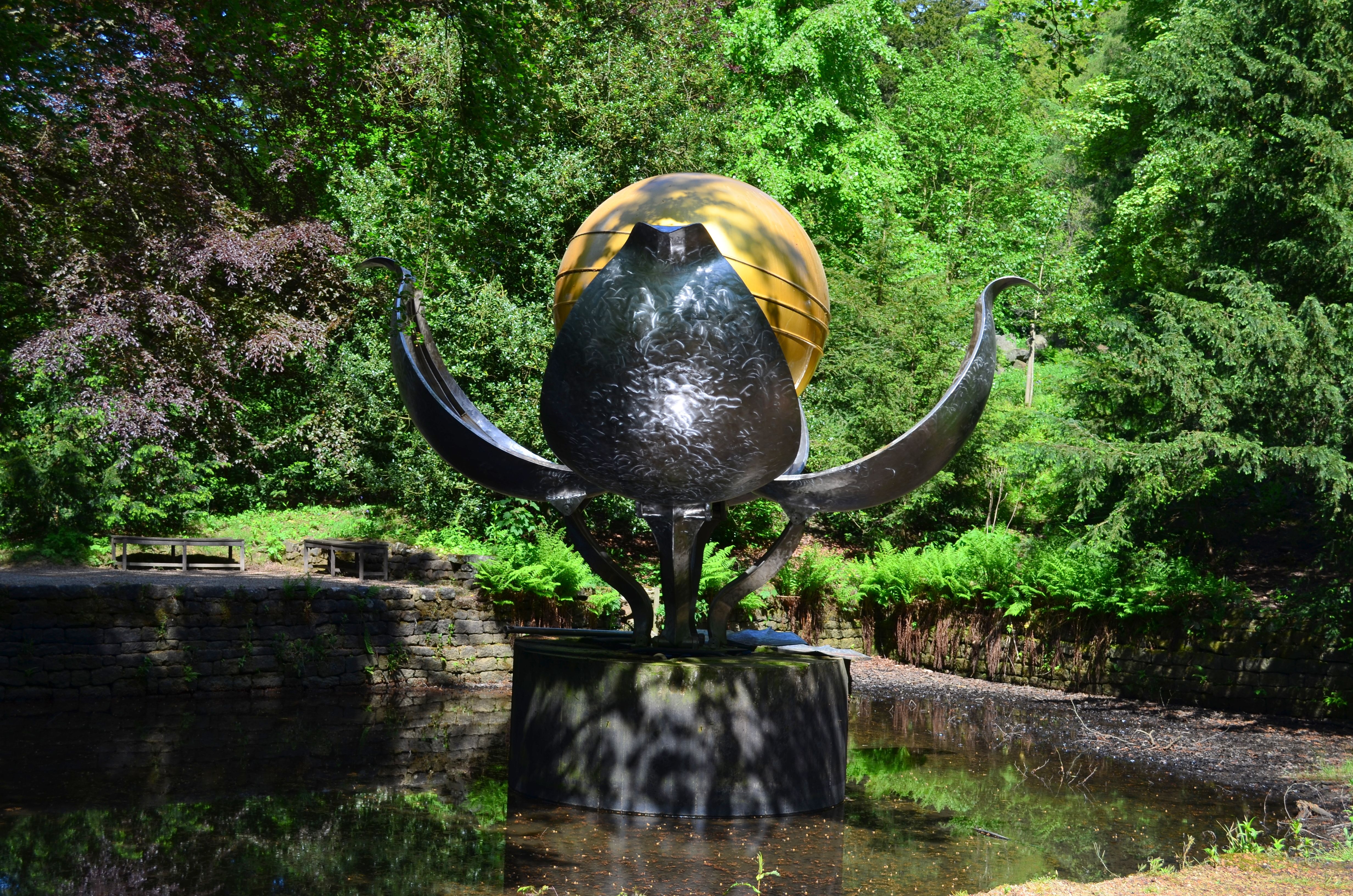 Artistic sculpture in a vibrant park surrounded by lush greenery and shadows reflecting in water.