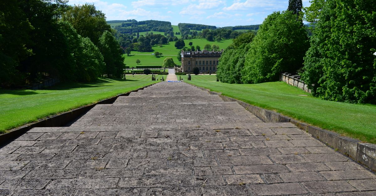 Wide angle view of Chatsworth House garden path, showcasing lush greenery and expansive landscape under a bright blue sky.