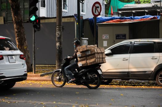 Motorcyclist carrying stacked boxes, waiting at a traffic light in a city street scene.