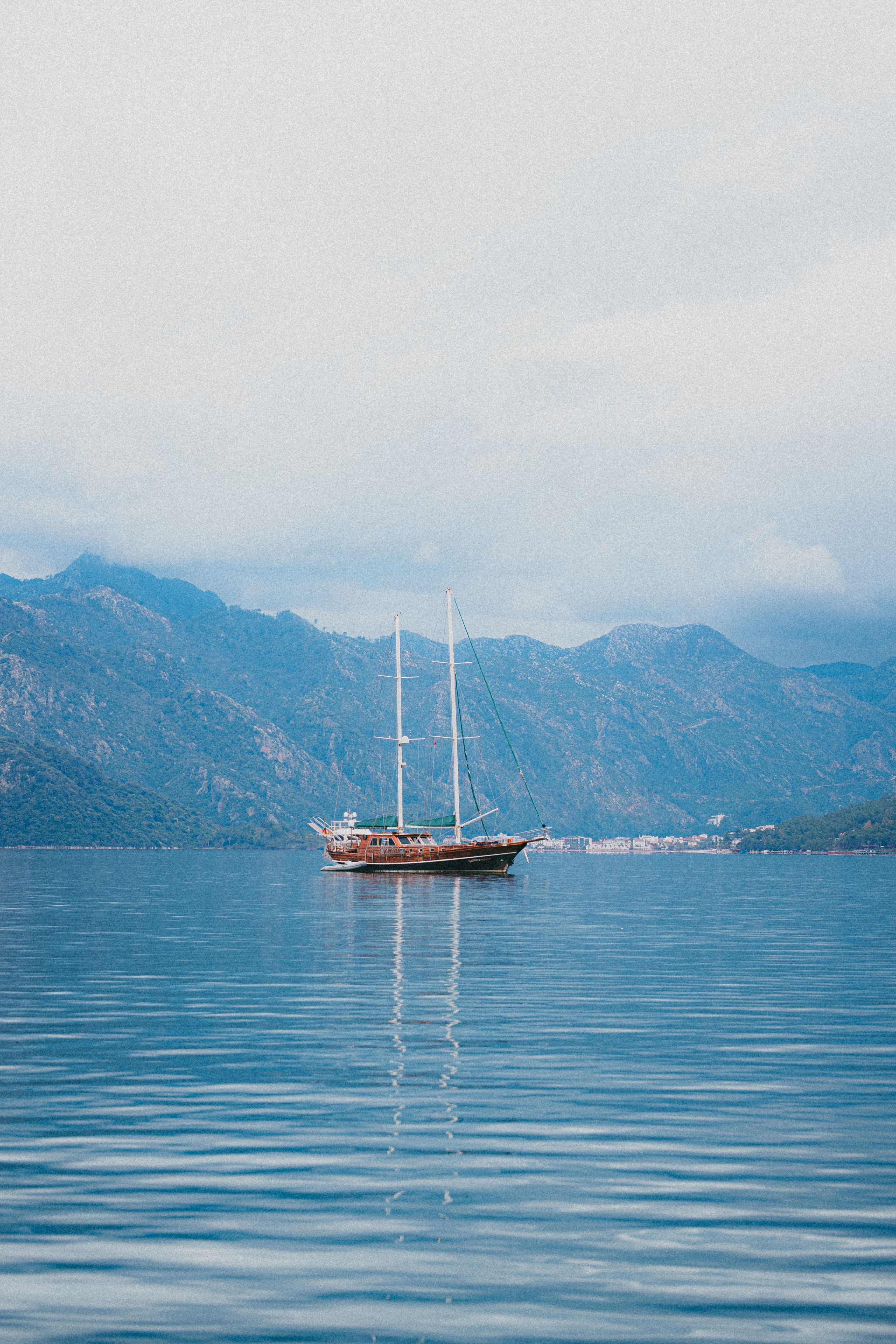 A tranquil sailing boat glides on a serene lake surrounded by towering mountains under a cloudy sky.