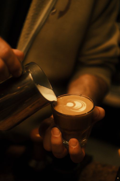 Free Close-up of barista pouring latte art into coffee cup in a warm cafe setting. Stock Photo