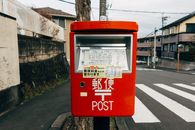 在日本小镇上，明亮的红色邮箱前，背景是人行横道和城市建筑