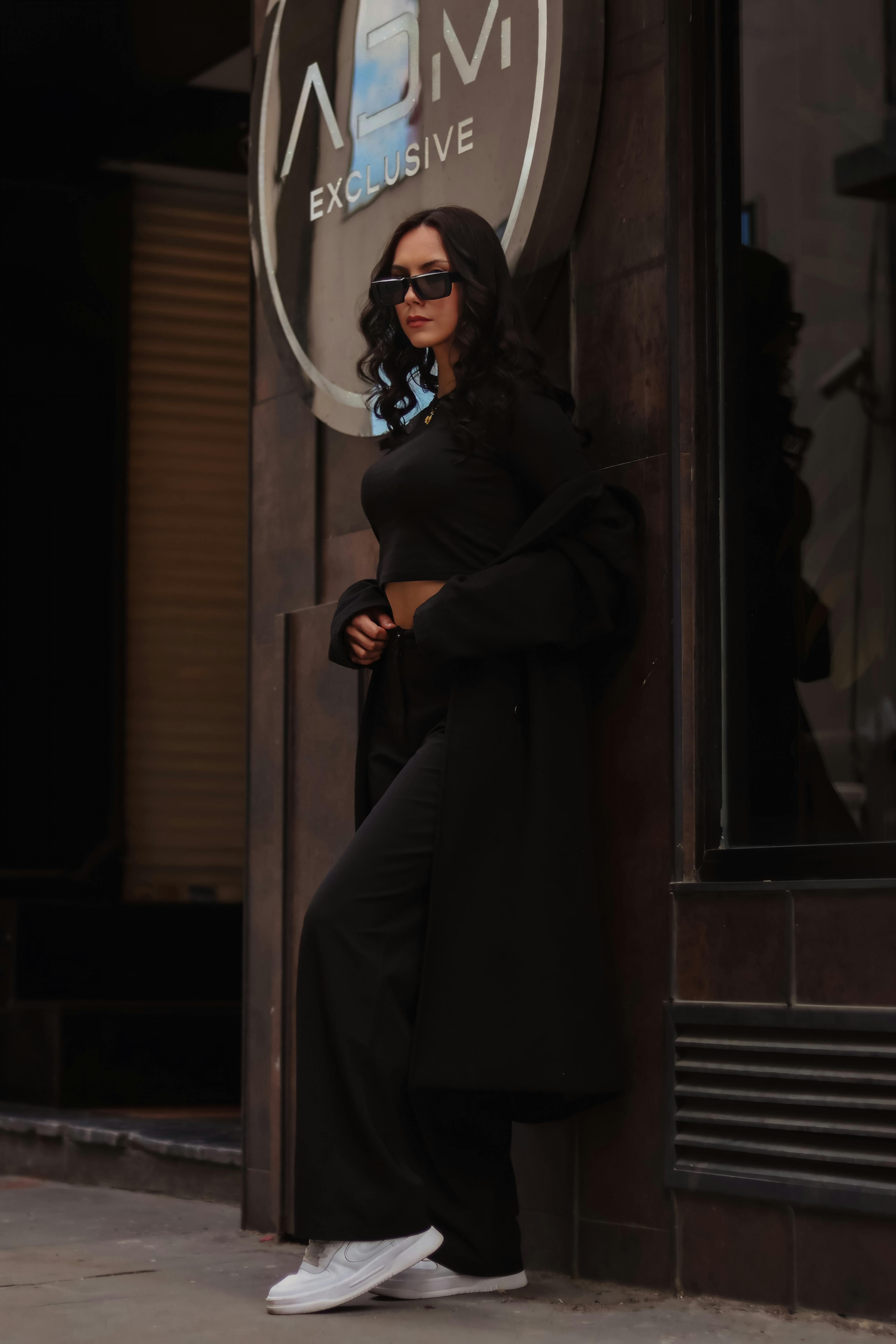 A fashionable woman in black attire poses confidently by a building exterior.