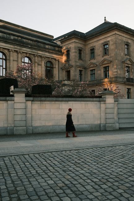 A lone figure walks by a historic building in Berlin, framed by blossoming trees.