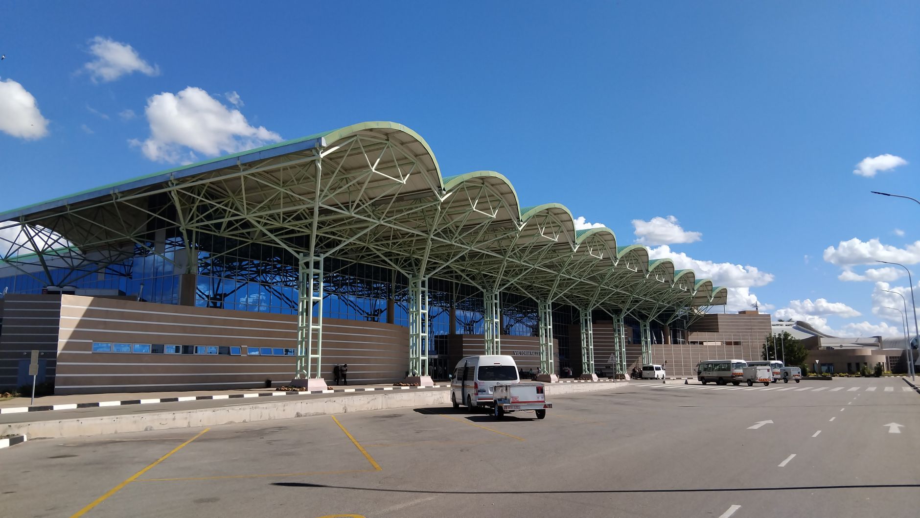Exterior view of a modern airport terminal with vehicles parked outside under a bright blue sky.