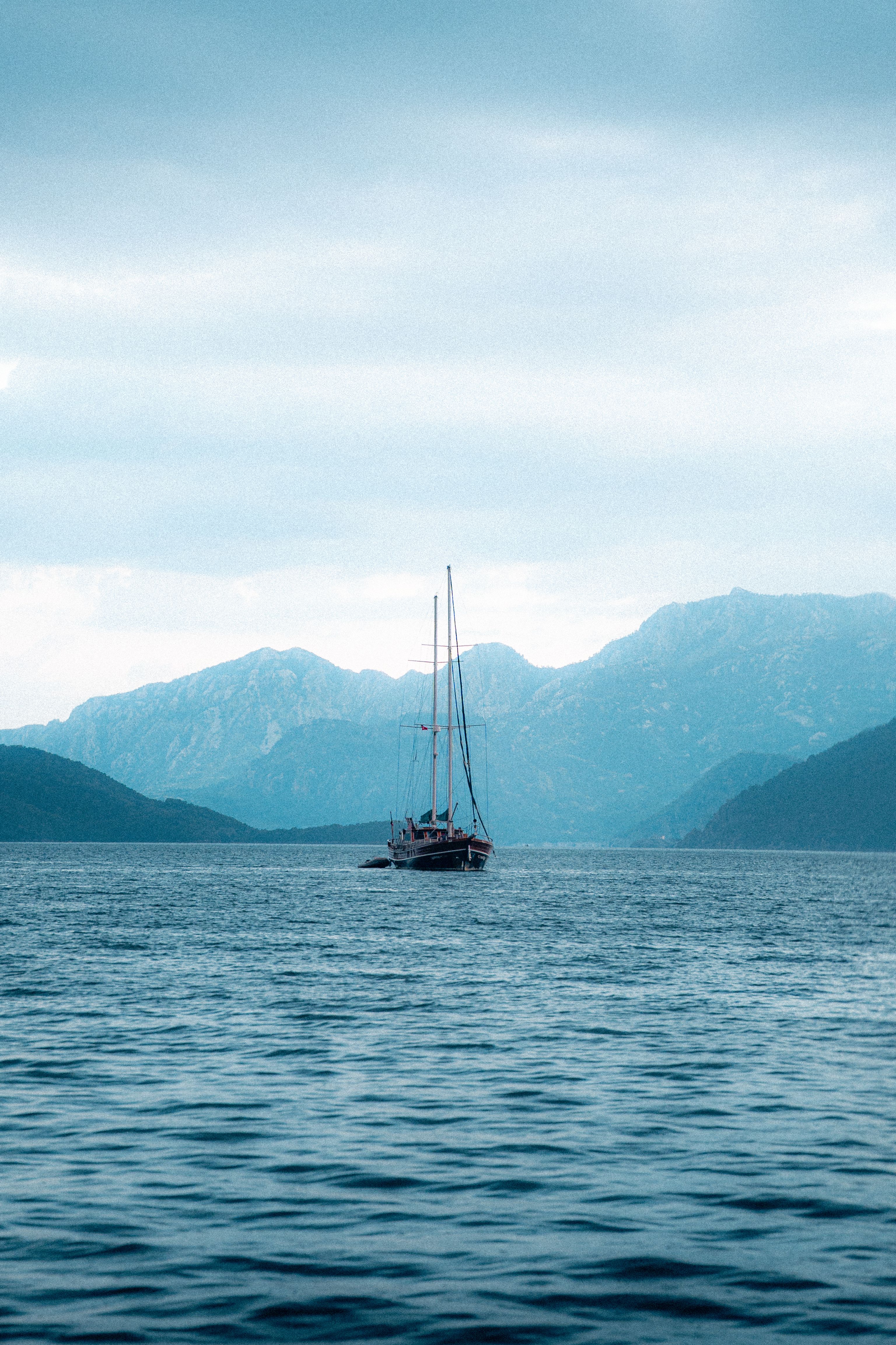 A solitary sailboat on a calm lake with a backdrop of majestic mountains under a cloudy sky.