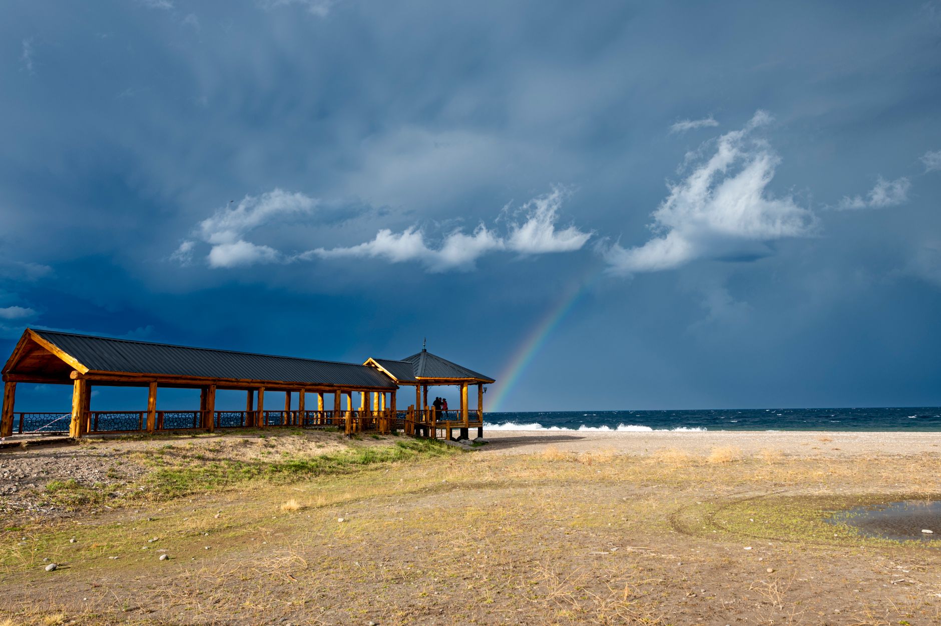 A wooden gazebo by the sea under dramatic skies with a rainbow in Santa Cruz, Argentina.