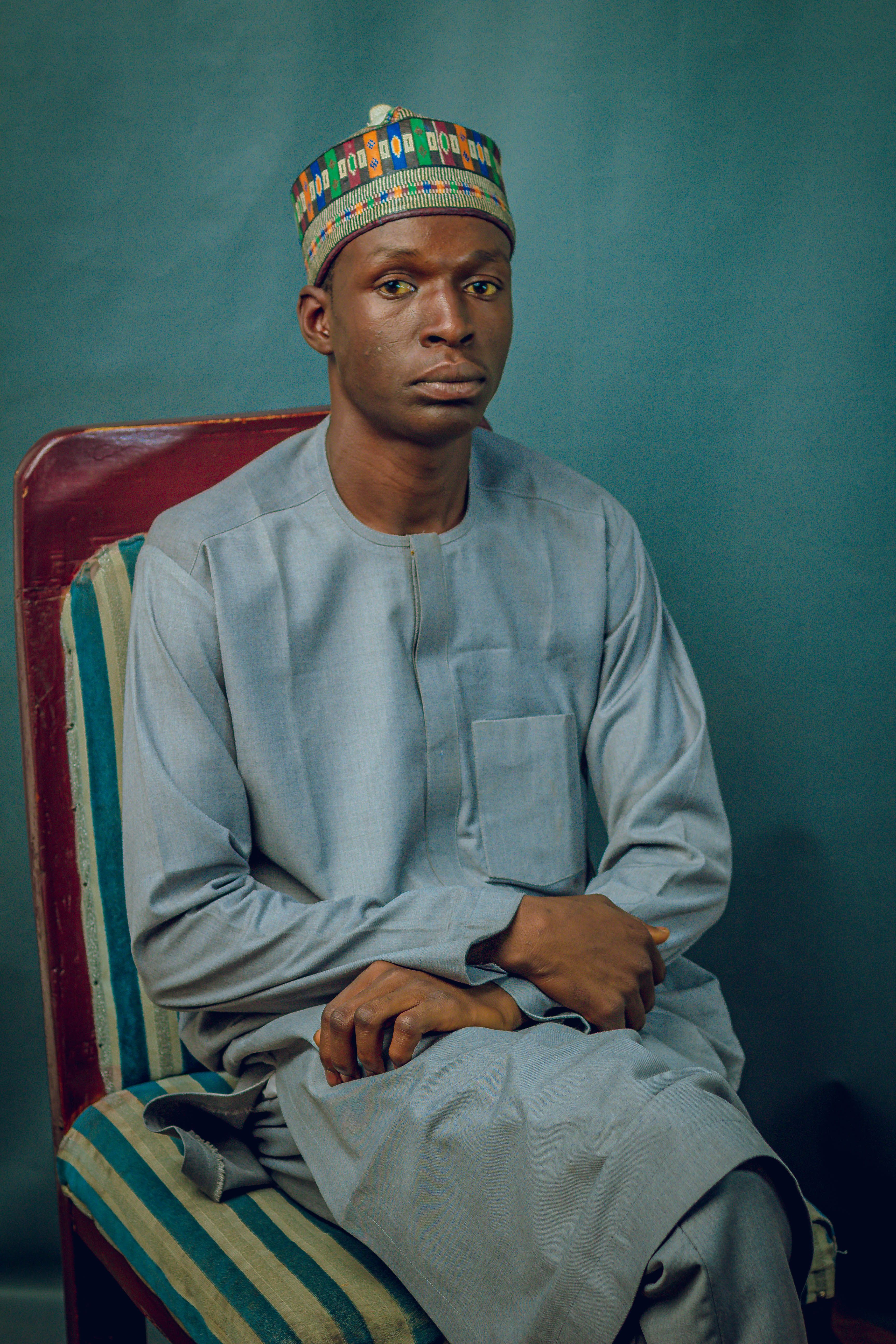 Man in traditional attire and colorful hat seated against a teal background.